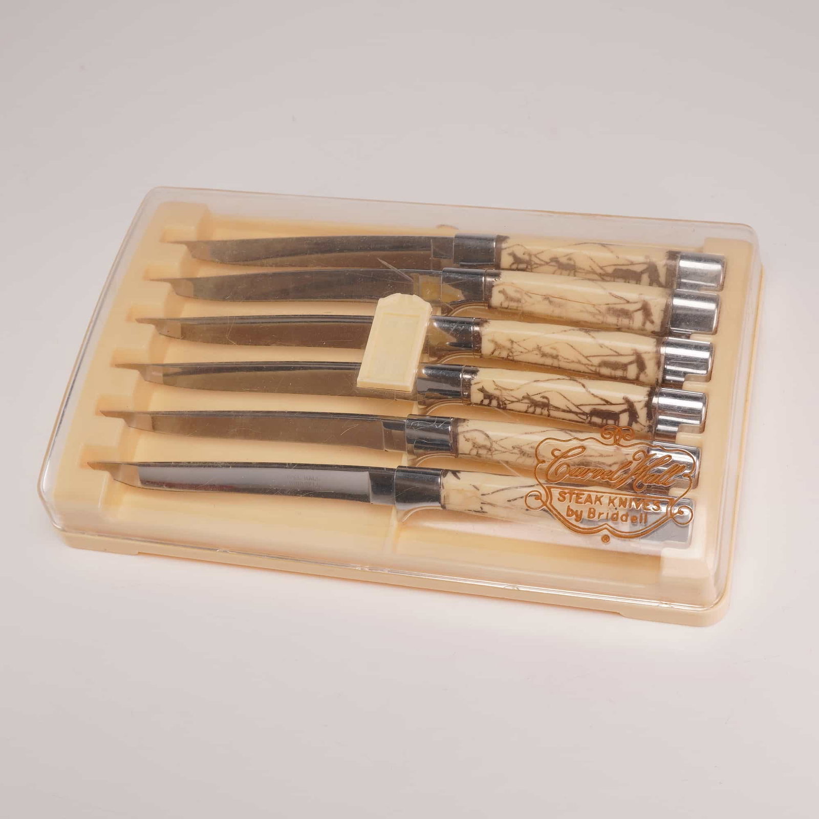 Set of Six Carvel Hall by Briddell Steak Knives: The set of six steak knives is marked Carvel Hall by Briddell on the stainless steel blades. The knives feature synthetic cream-colored handles with dog sled design and polished metal bolsters and end