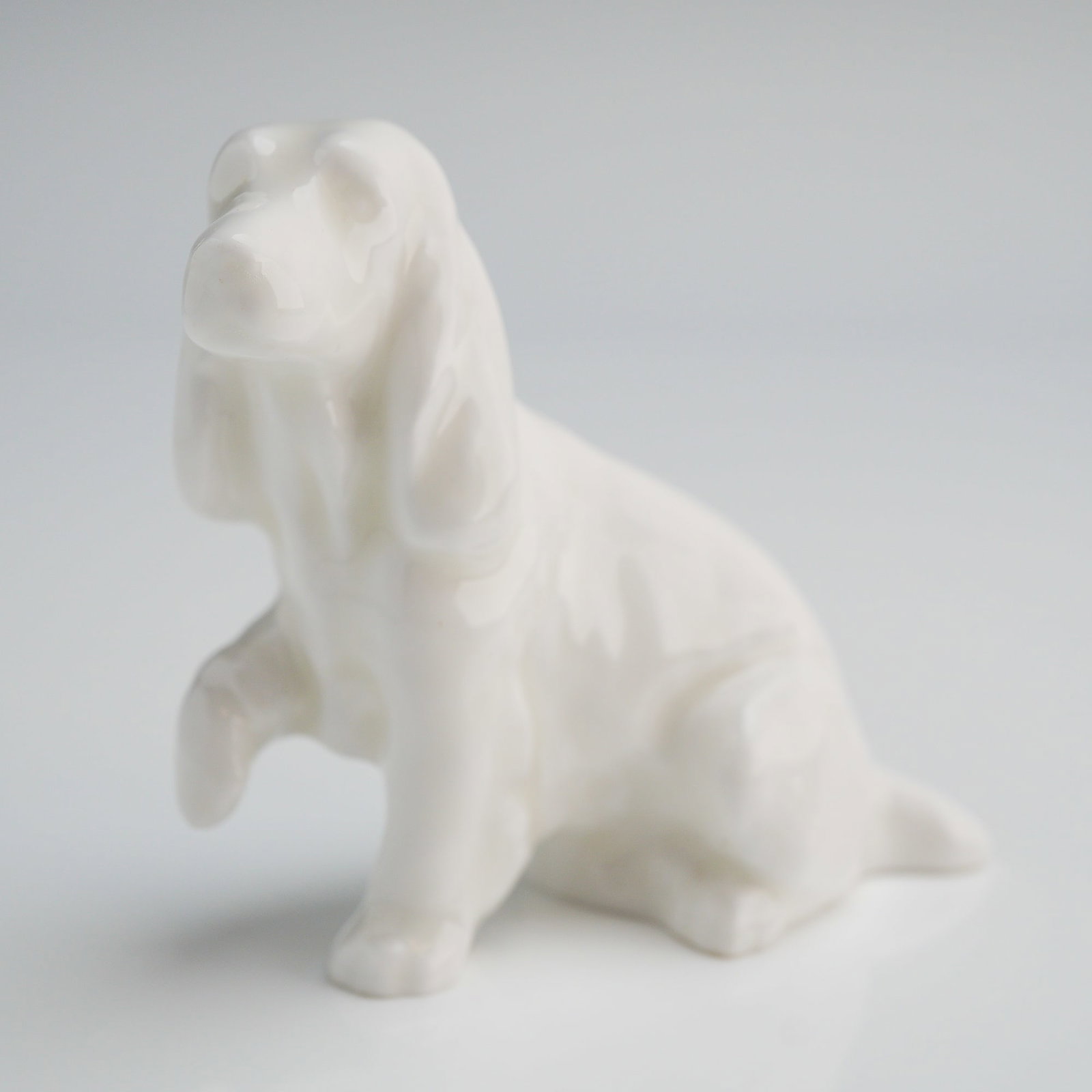 Royal Doulton Bone China Cockder Spaniel Dog K9A in Rare White Colourway: A single ceramic dog figurine crafted from highly glazed white bone china. The object is depicted in a sitting pose with one forepaw raised, consistent with a spaniel breed. The style is traditional, 