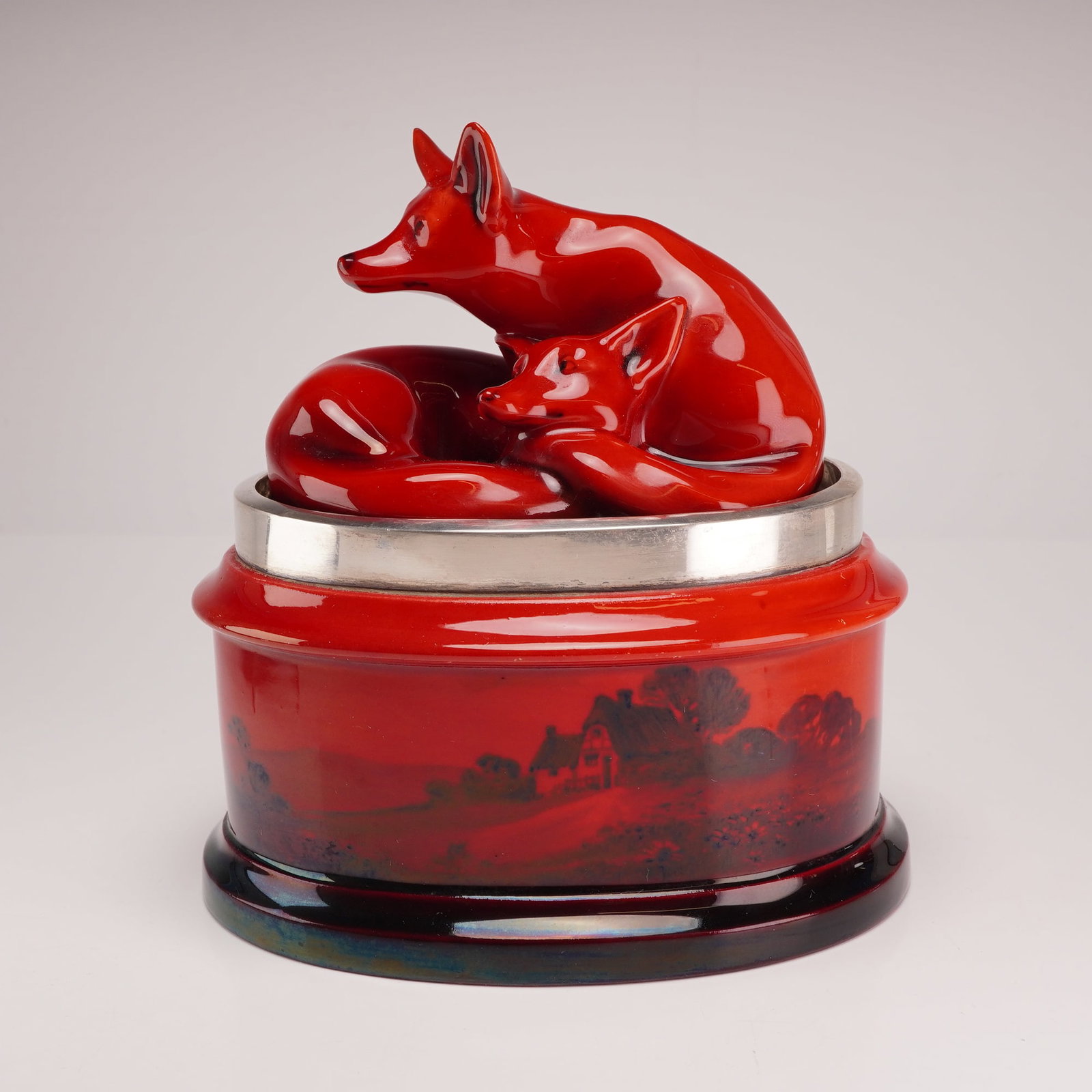 Royal Doulton Flambe Lidded Tobacco Jar with Sterling Silver Rim, Curled Foxes: A Royal Doulton Flambe ceramic tobacco jar featuring a figural lid and a hallmarked silver rim. The lid is topped with a finial depicting two foxes, one seated in an alert posture and the other curled
