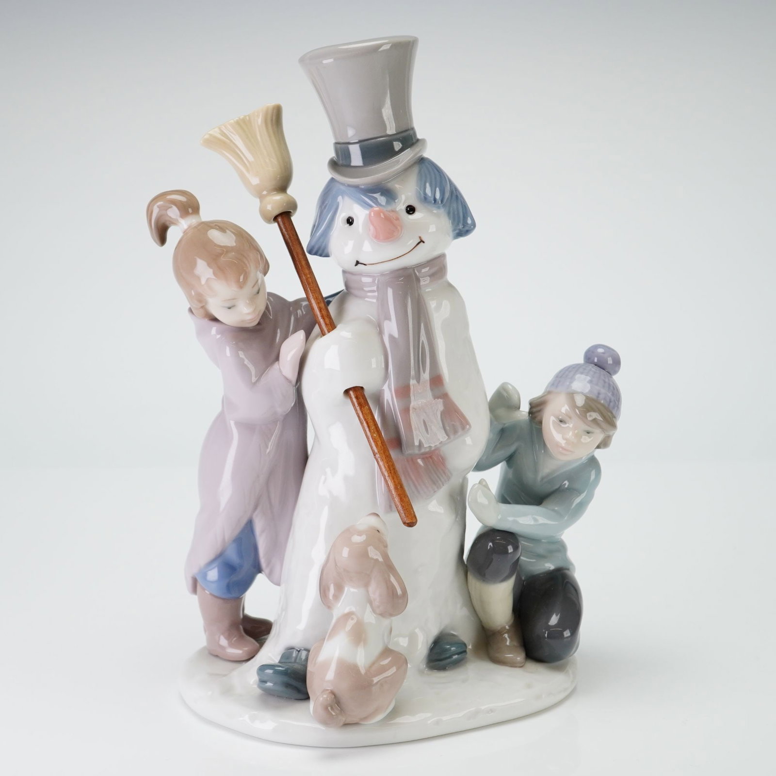 Lladro Porcelain Figurine, The Snow Man 1005713: The scene depicts a young girl in a lavender coat and a boy in a blue jacket and knit cap placing a broom into the hand of a snowman. The snowman is further detailed with a grey top hat and a striped
