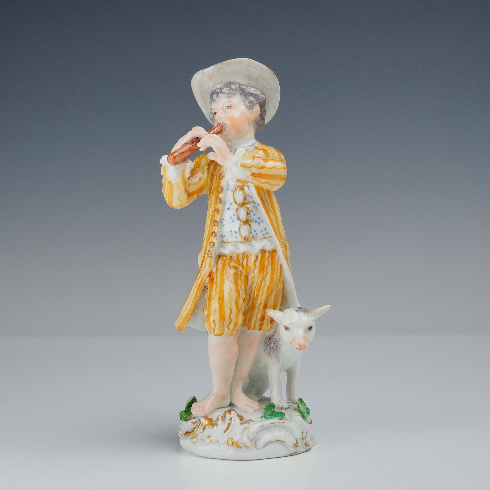 Meissen Porcelain Figurine Young Boy Playing a Flute with Dog at His Feet: This Meissen porcelain figurine depicts a young boy shepherd in 18th-century attire playing a transverse flute. The figure is dressed in a yellow and orange striped frock coat with matching breeches,