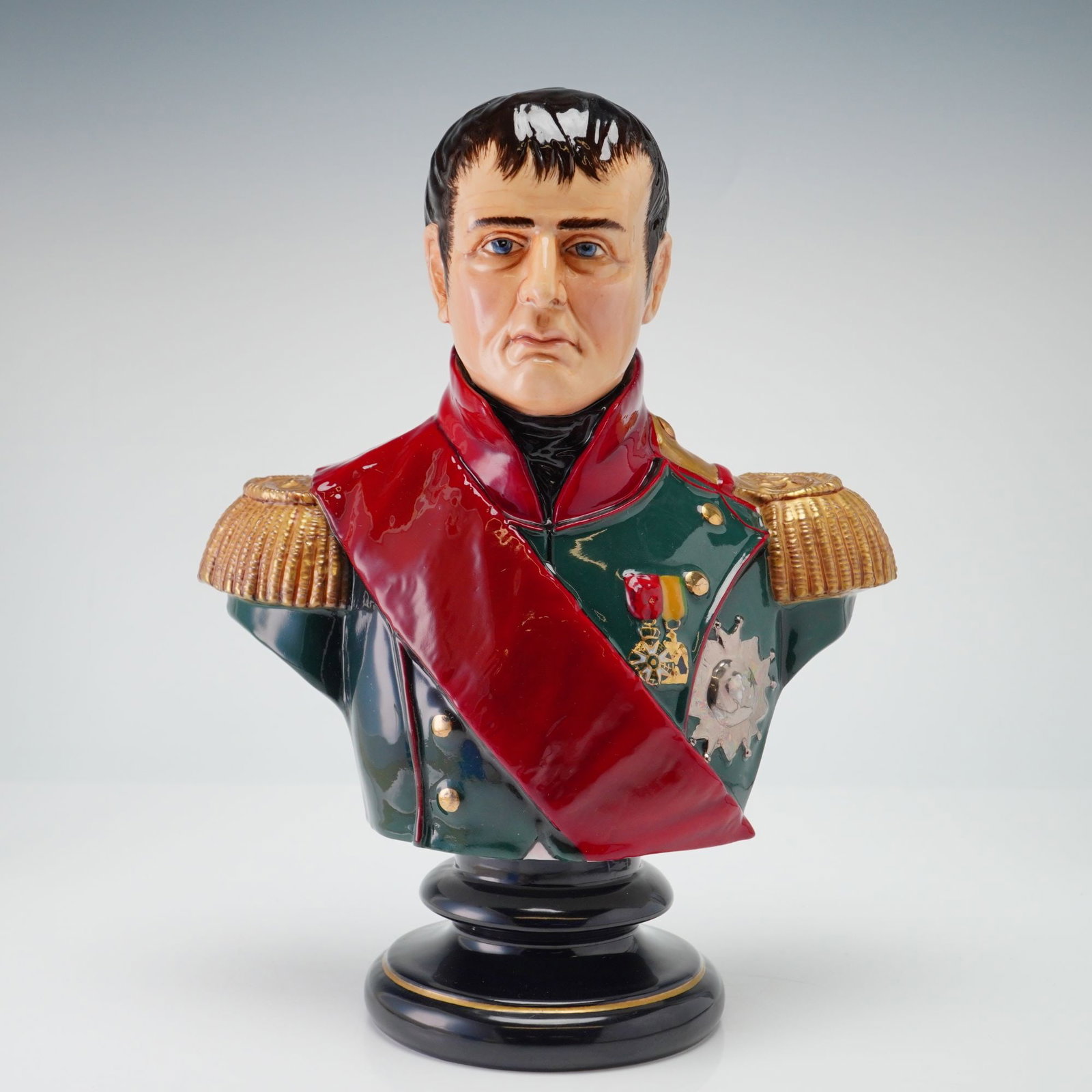 Michael Sutty Fine China Bust, Napoleon Bonaparte 1815: This Michael Sutty porcelain portrait bust depicts Napoleon Bonaparte as he appeared in 1815. The figure is rendered in polychrome glazes, wearing a green military tunic with a red collar and a red sa