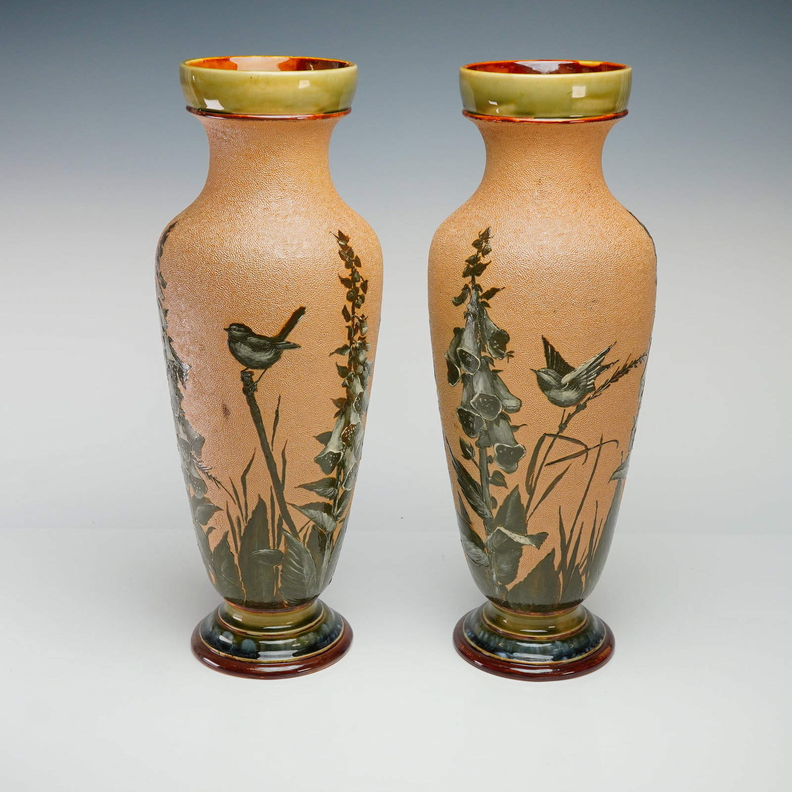 Pair of Doulton Lambeth by Florence Barlow Stoneware Vases: Two English stoneware vases with textured grounds, each decorated with a pate-sur-pate scene of birds perched among foxglove flowers. The feet finished in glossy green, gray, and brown glazes, and the