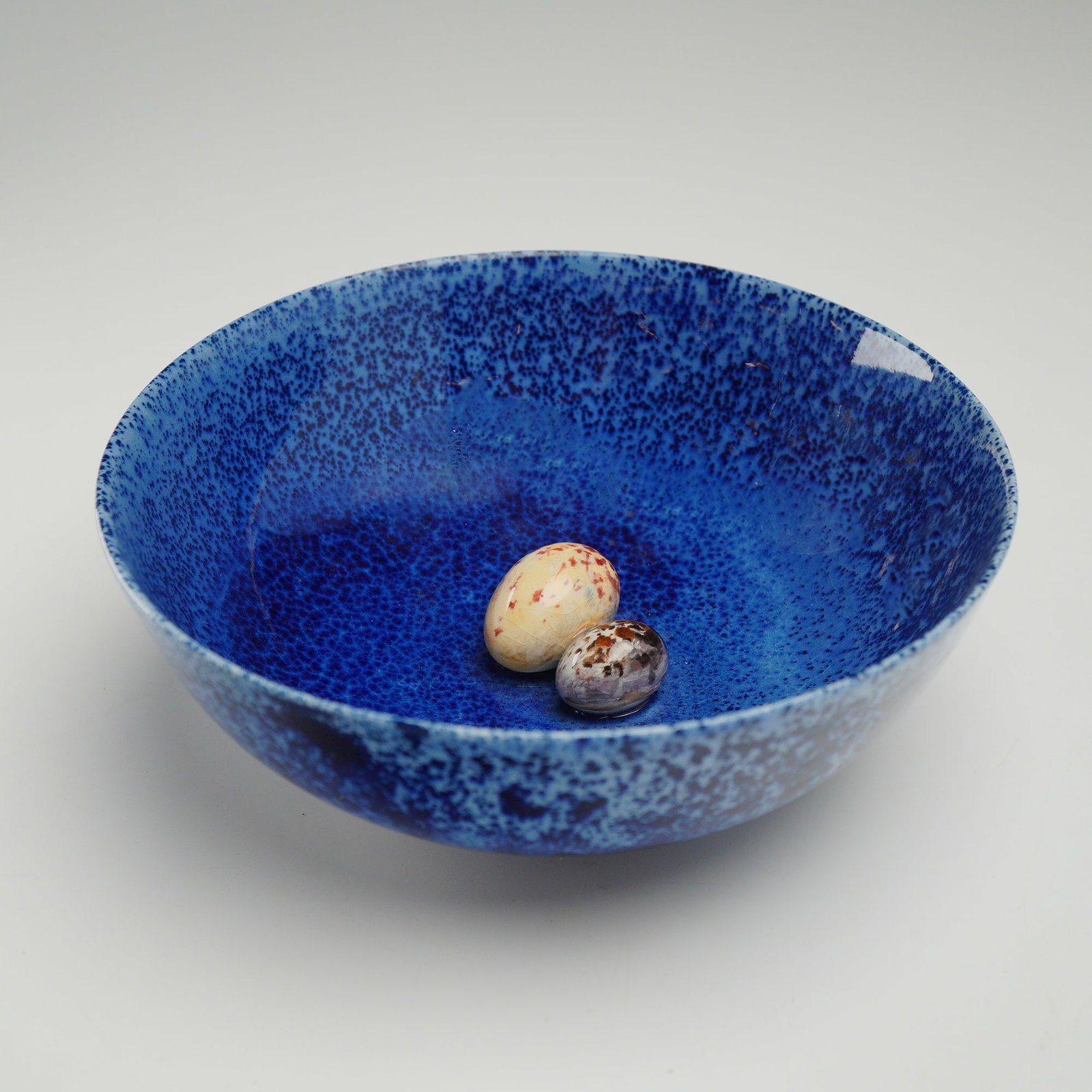 Royal Doulton Titanian Speckled Blue Bowl with Two Ceramic Eggs by J. Birbeck Senior: This Royal Doulton Titanian ware bowl is finished in a characteristic speckled and mottled cobalt blue glaze. The lot includes two solid ceramic eggs intended for display within the bowl, each glazed
