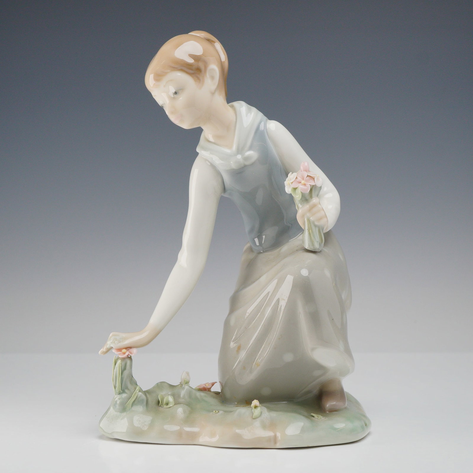Lladro Porcelain Figurine Girl Gathering Flowers 1001172: Features a young girl kneeling on a grassy base while gathering handmade pink flowers, dressed in a blue tunic and tan skirt. The base displays the blue stamped factory mark LLADR" / HAND MADE IN SPAI