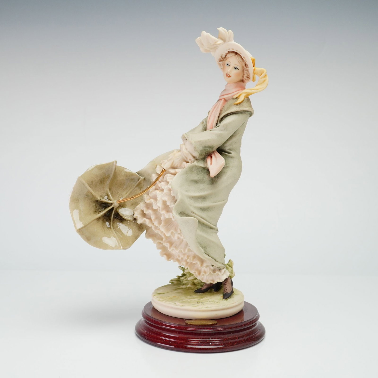 Giuseppe Armani Florence Figurine Stormy Weather 0533-C: The piece is crafted from cold-cast porcelain with a matte finish and depicts a woman in early 20th-century attire, featuring a long olive-green coat, a voluminous tiered ruffled skirt, a pink scarf, 