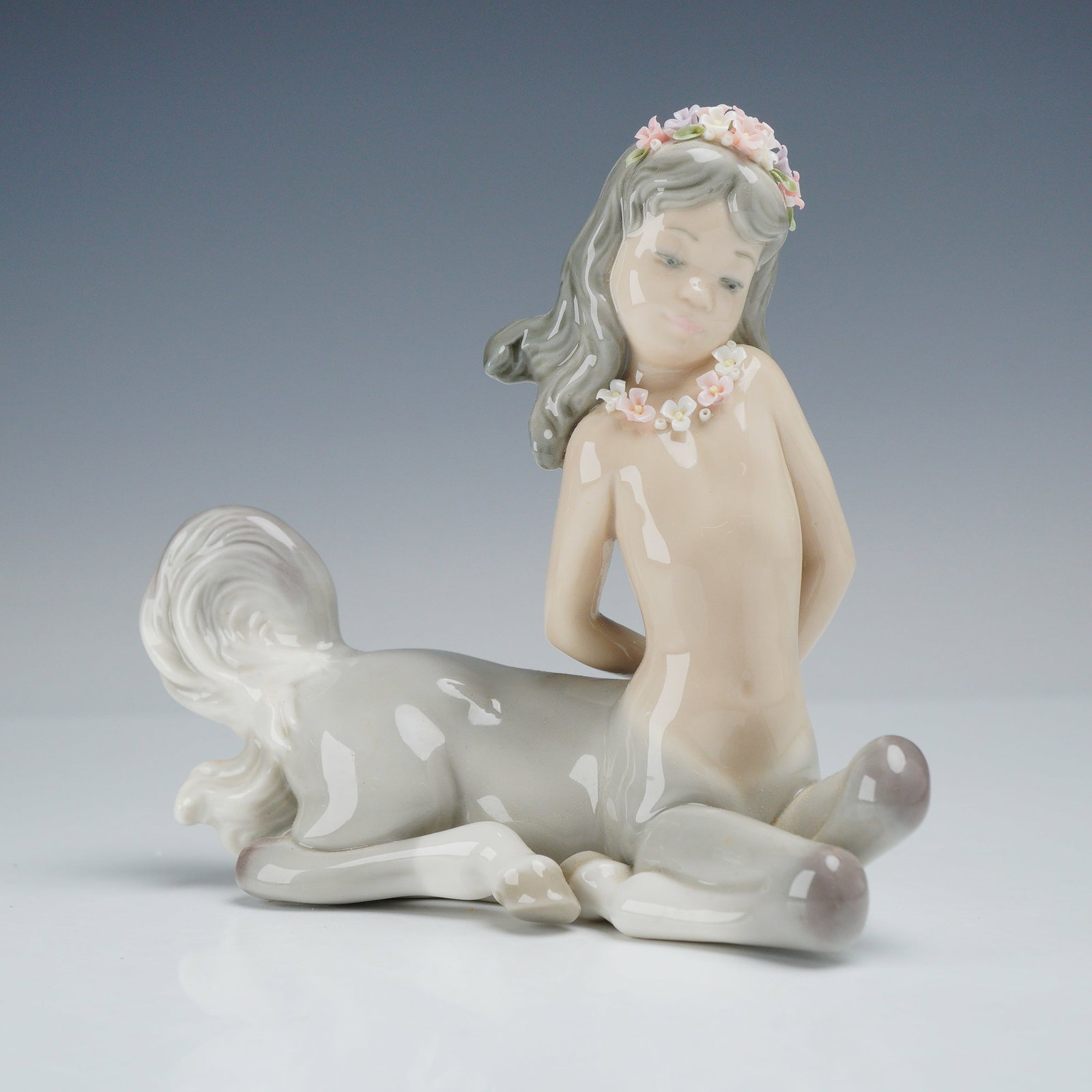 Lladro Porcelain Figurine, Demure Centaur Girl 01005320: This glazed porcelain figurine, produced by Lladro, depicts a reclining female centaur, often referred to as a centaurette. The figure features a soft grey equine lower body and a tan human torso. The