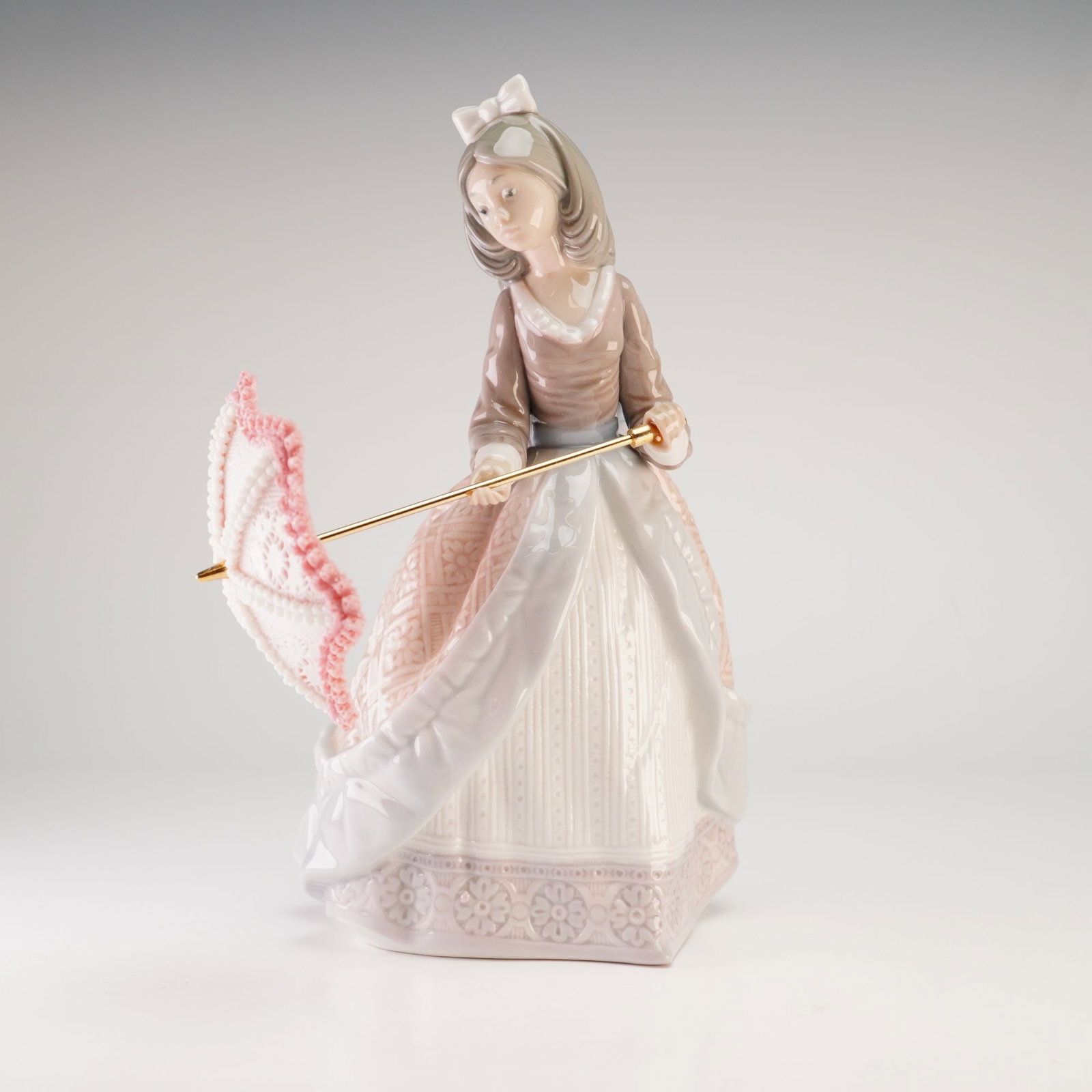 Lladro Porcelain Figurine, Jolie 1005210: The figurine depicts a young woman in 19th-century attire, featuring a long, tiered gown in shades of mauve, white, and pale pink. The dress is accented with a large light blue bow at the back of the 