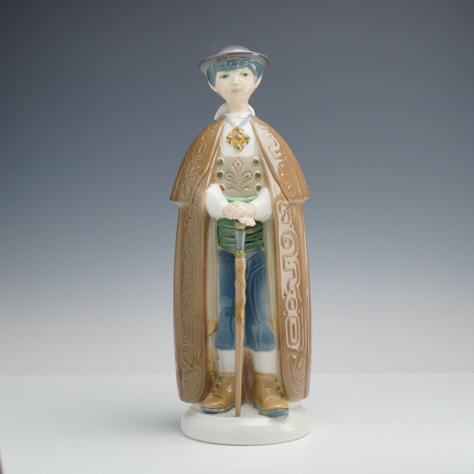 Lladro Porcelain Figurine, Little Town Mayor 1001161: Depicts a standing male figure in traditional Spanish regional attire. The figure is dressed in a long, tan-colored cloak featuring relief-molded geometric and floral patterns. Underneath the cloak, h