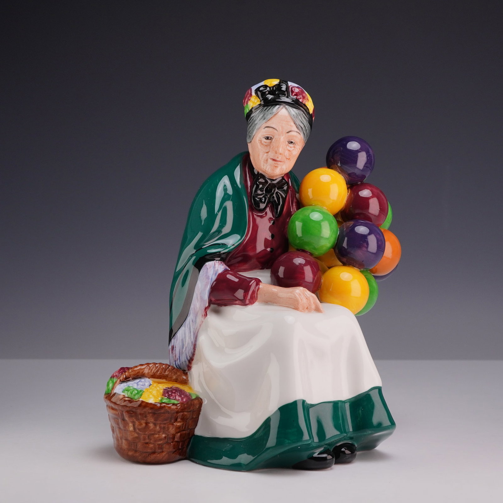 Royal Doulton Figurine, Old Balloon Seller HN1315: The figure depicts an elderly woman seated on a brown crate. She is dressed in a green shawl over a red bodice and a white apron with a dark green border, with a patterned headscarf tied over her grey