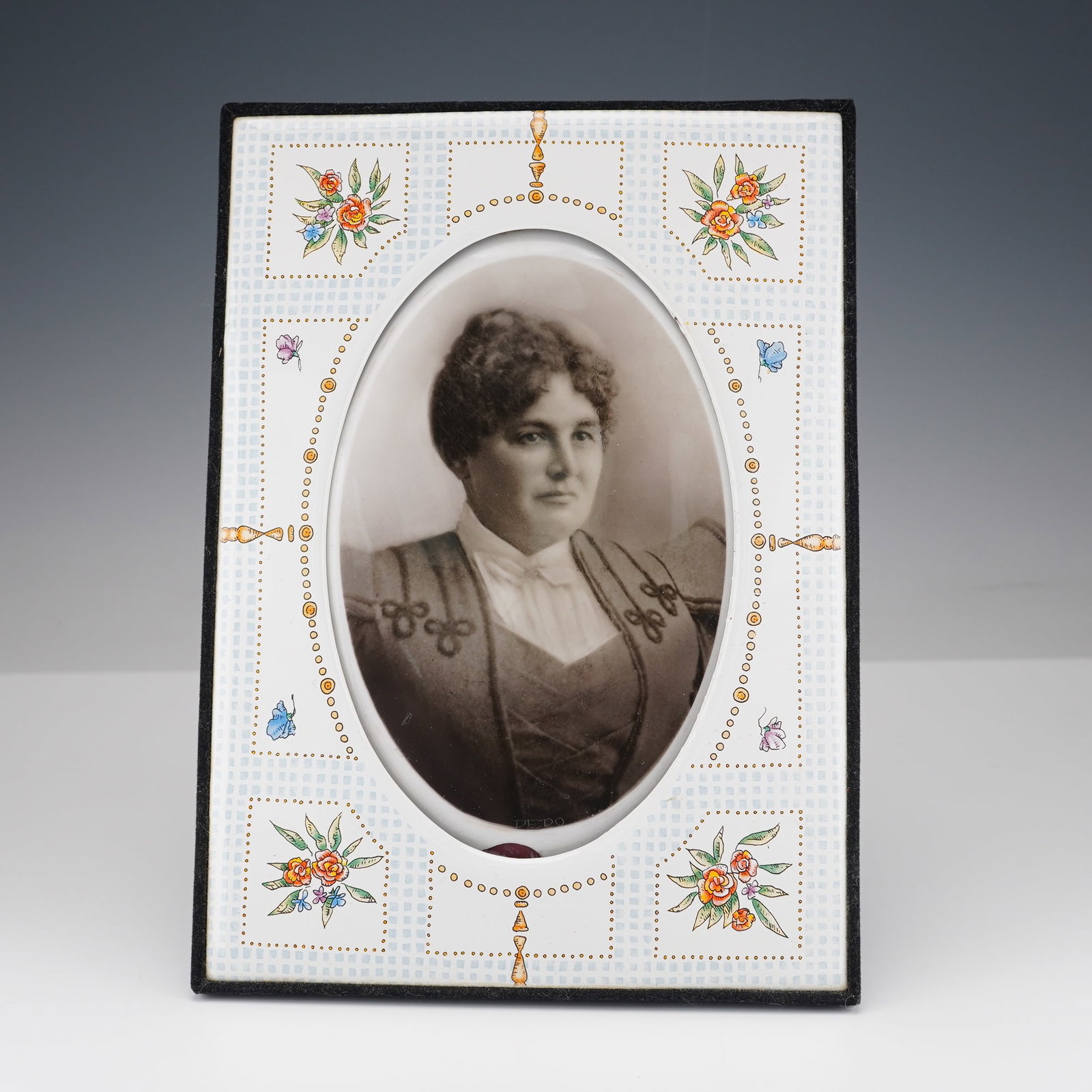 Enamel Portrait Frame with Antique Photograph on Convex Enamel Base: A refined enamel portrait frame showcasing a beautifully detailed oval photograph of a woman, with Dedo written on the lower edge. The frame is adorned with delicate hand-painted floral motifs in shad