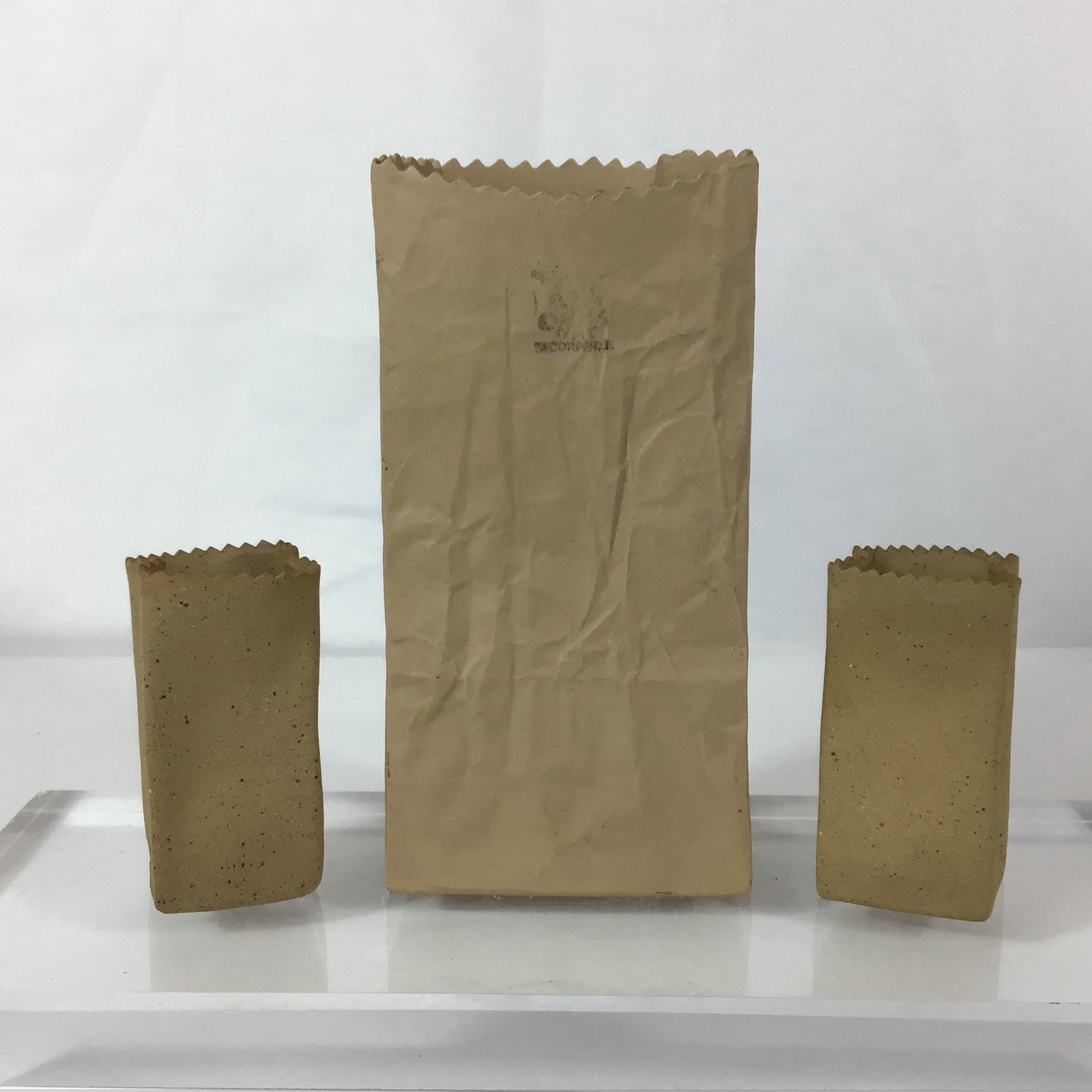 3pc Michel Harvey Ceramic Brown Paper Bag Sculptures: A trio of tan ceramic sculptures designed in the trompe-l'"il style of brown paper bags, featuring one larger central bag with a faint circular mark and two smaller flanking bags. Dimensions: 3.00"L x