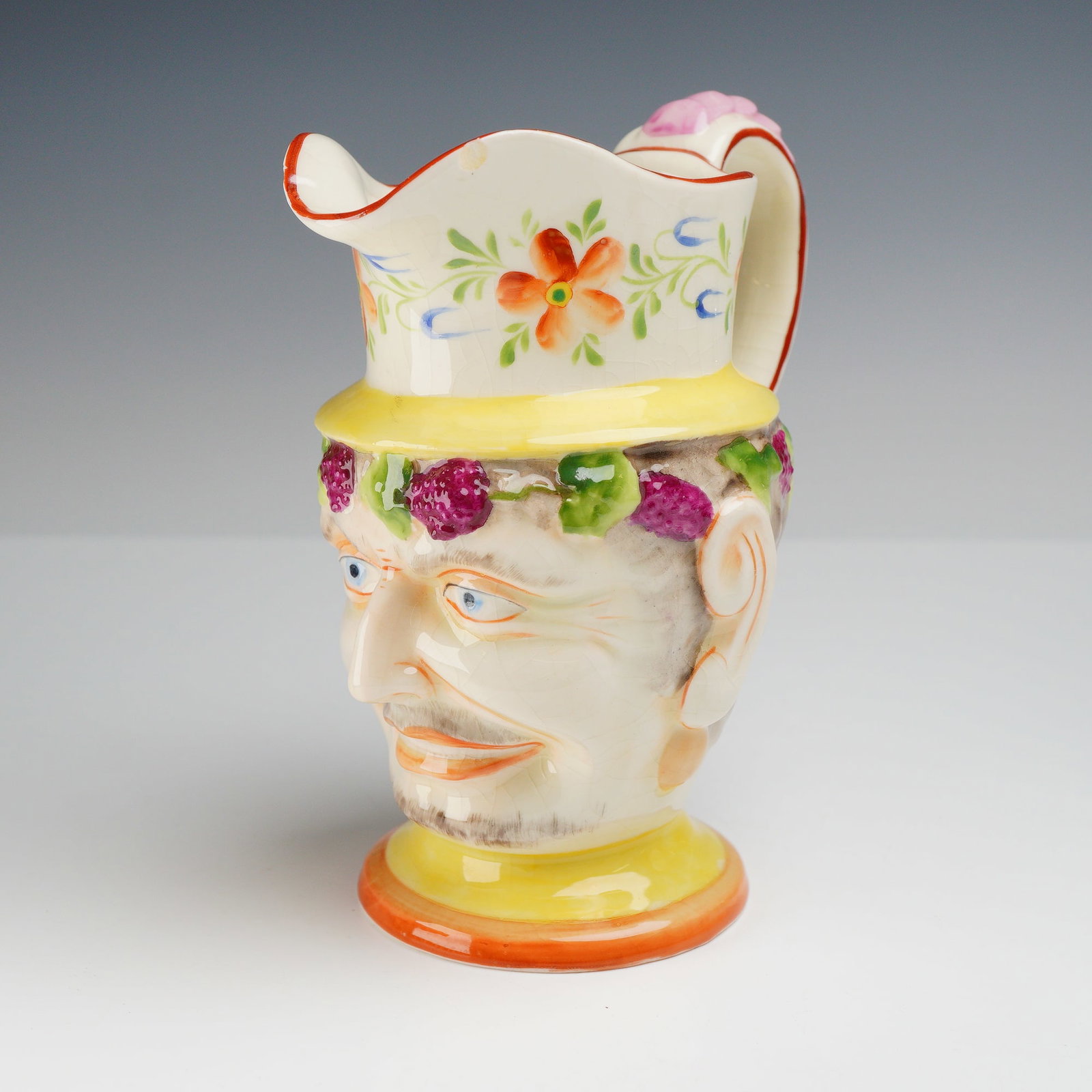 Staffordshire Pearlware Satyr Mask Jug: This ceramic jug is modeled as a satyr or Bacchus mask, a form attributed to Staffordshire potteries in the early 19th century. The vessel is constructed of earthenware, likely pearlware, featuring re