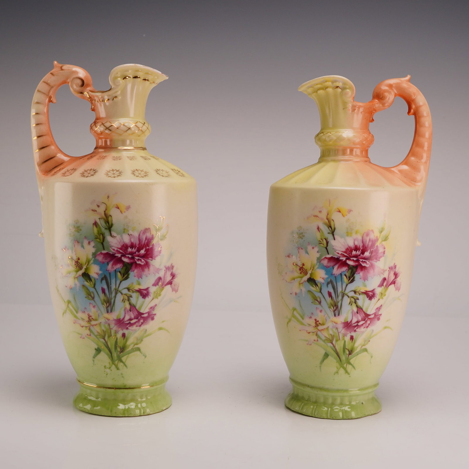 Pair of Royal Floretta Ware Hand Painted Floral Pitchers: Each pitcher features a slender ovoid body tapering to a pedestal base. The narrow neck is accented with a bulbous, textured ring below a flared, scalloped spout. The ornate, high-arched handles featu