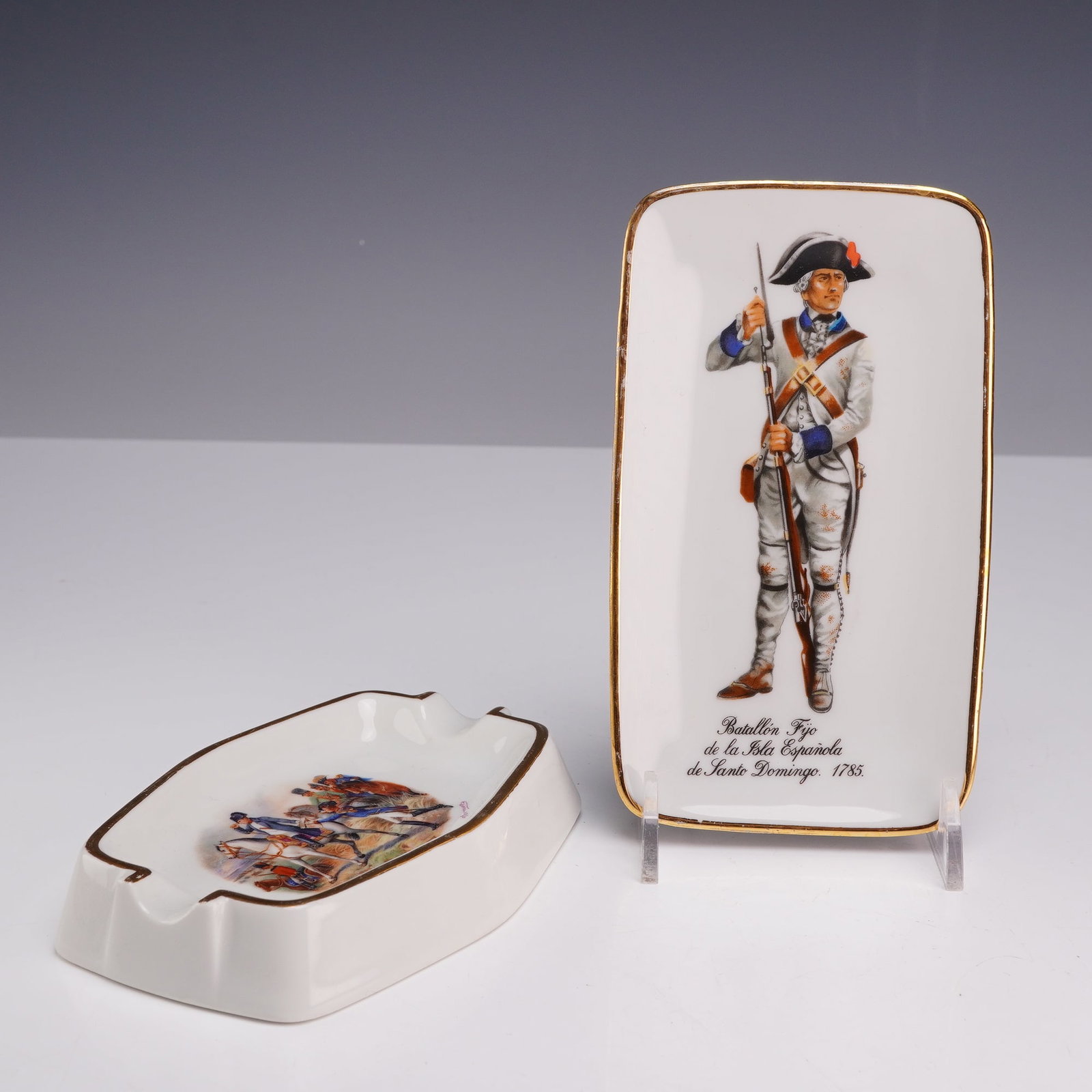Two Limoges Gilded Porcelain Military Themed Decorative Items: This lot consists of two pieces of French porcelain featuring historical military motifs. The collection includes one oblong ashtray and one rectangular pin tray, both constructed from white glazed po