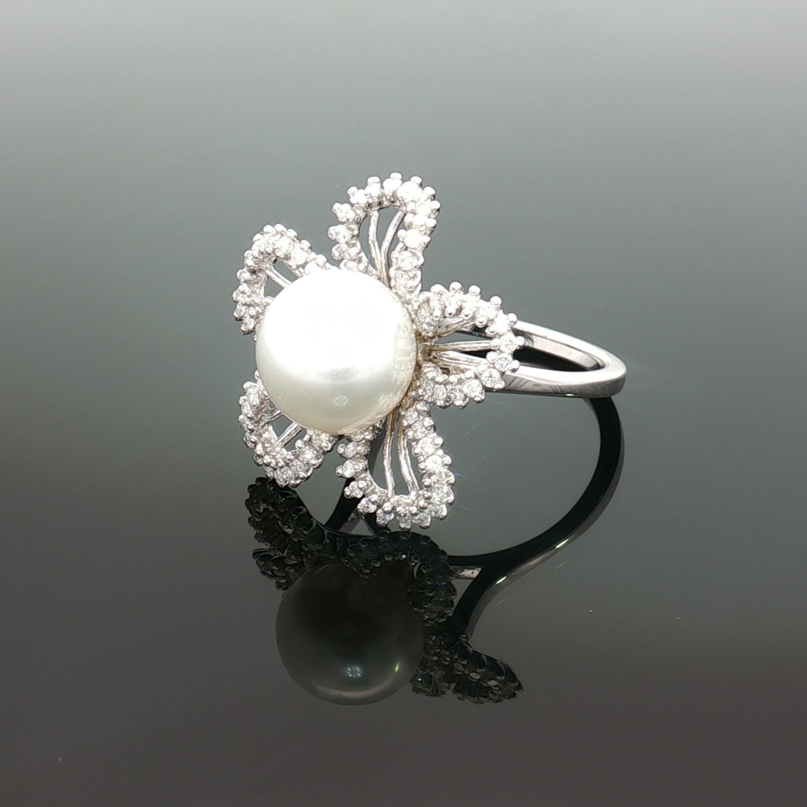 Estate 18K White Gold South Sea Pearl and Diamond Ring: An estate cocktail ring featuring a prominent round white South Sea pearl centerpiece framed by a five-petaled floral motif crafted in polished 18K white gold. Each petal features an open-work design 