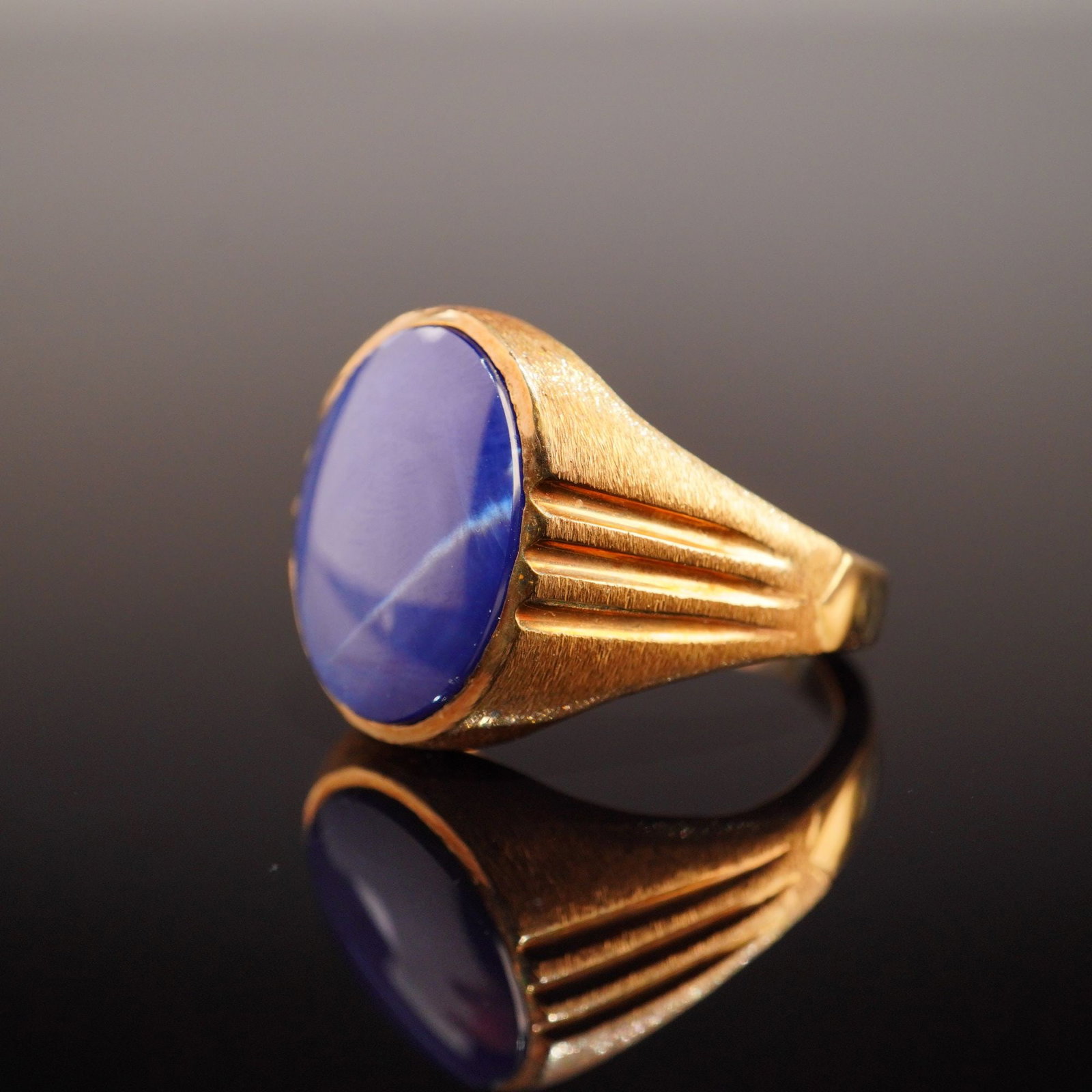 Men's 14k Yellow Gold Blue Star Sapphire Cocktail Ring: This men's signet-style cocktail ring is constructed of 14 karat yellow gold. The center is set with one oval cabochon-cut blue star sapphire in a protective bezel setting. The gemstone displays a pro