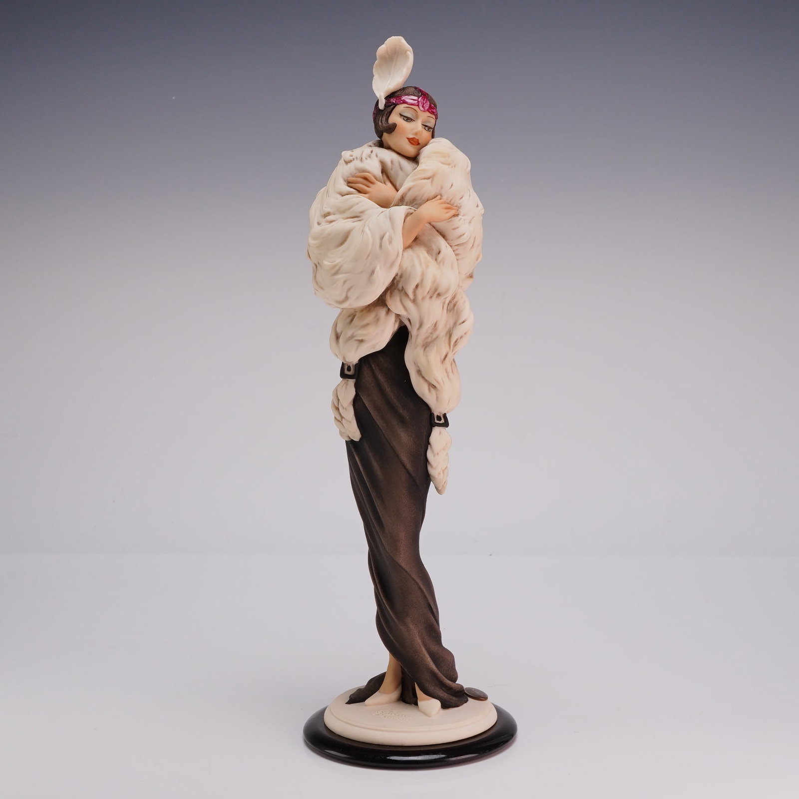 Giuseppe Armani Florence Figurine Gwendoline 1728C: The piece is rendered in an Art Deco style, featuring a woman in a floor-length dark brown gown and a voluminous, textured cream-colored fur stole. She wears a magenta-toned jeweled headband accented 