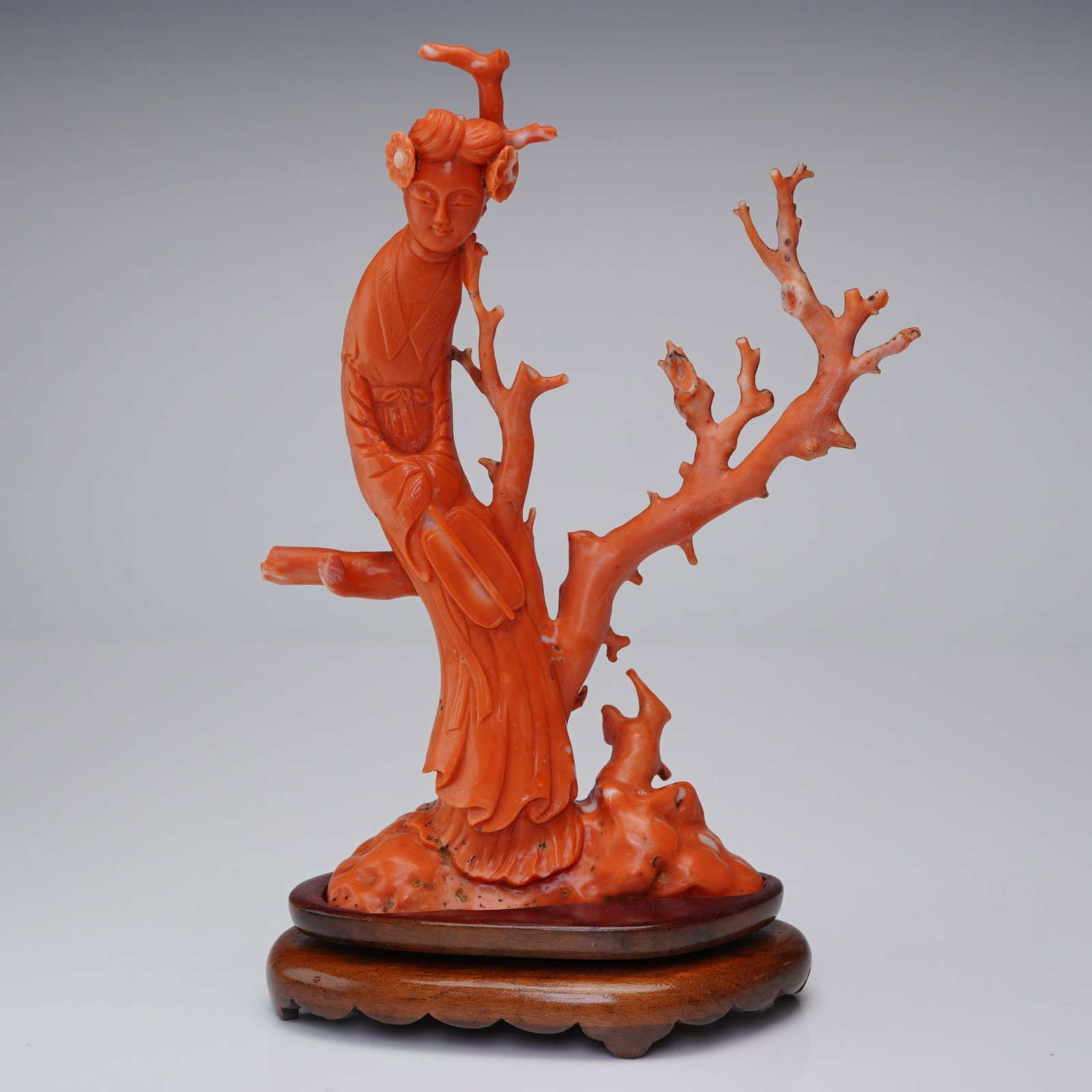 Chinese Carved Red Coral Figurine with Wooden Stand: The sculpture depicts a graceful maiden standing amidst a network of gnarled, budding branches. She is dressed in traditional flowing robes featuring finely incised key-fret patterns at the collar and