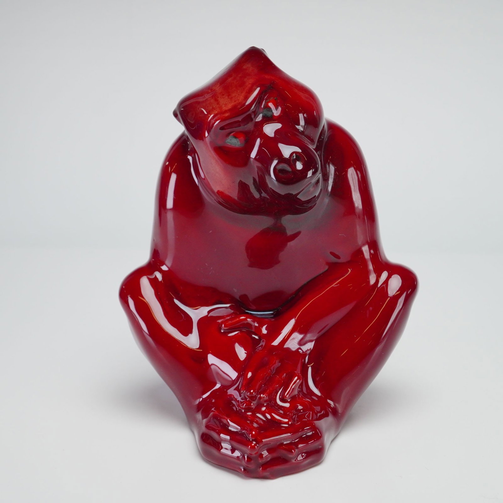 Royal Doulton Experimental Flambe Figurine, Ape HN140: Depicting a seated ape in a contemplative pose with hands clasped over its feet. The figure is finished in a rich, high gloss flambe red glaze, featuring deep, varied red tones characteristic of a red
