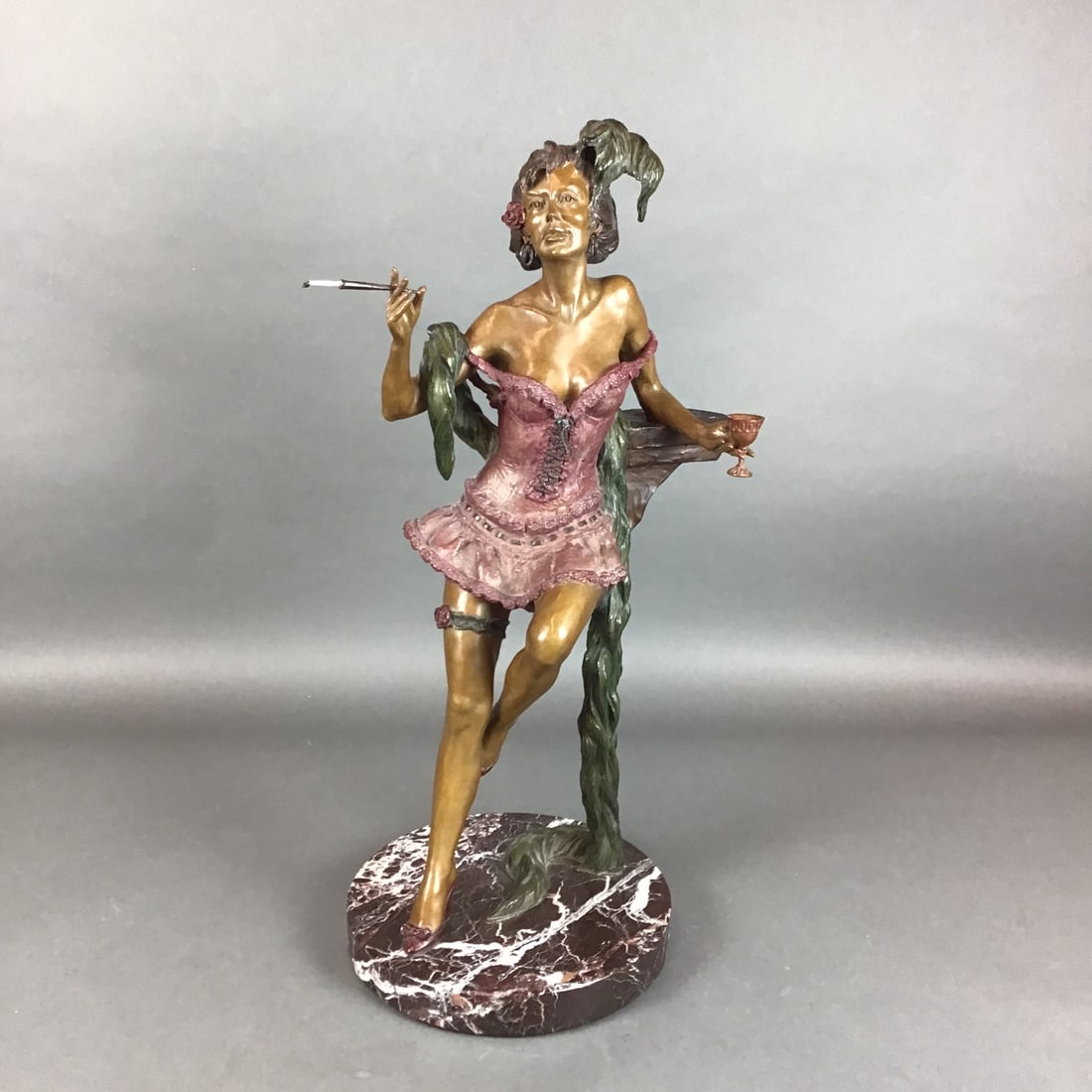 Eli Hazak (Israeli-American, b. 1945) Bronze Sculpture, Calico Queen, Signed: This contemporary polychrome bronze sculpture by artist Eli Hazak depicts a 19th-century style saloon girl. The figure is dressed in a pink corset with ruffled trim and a feathered headpiece, leaning 