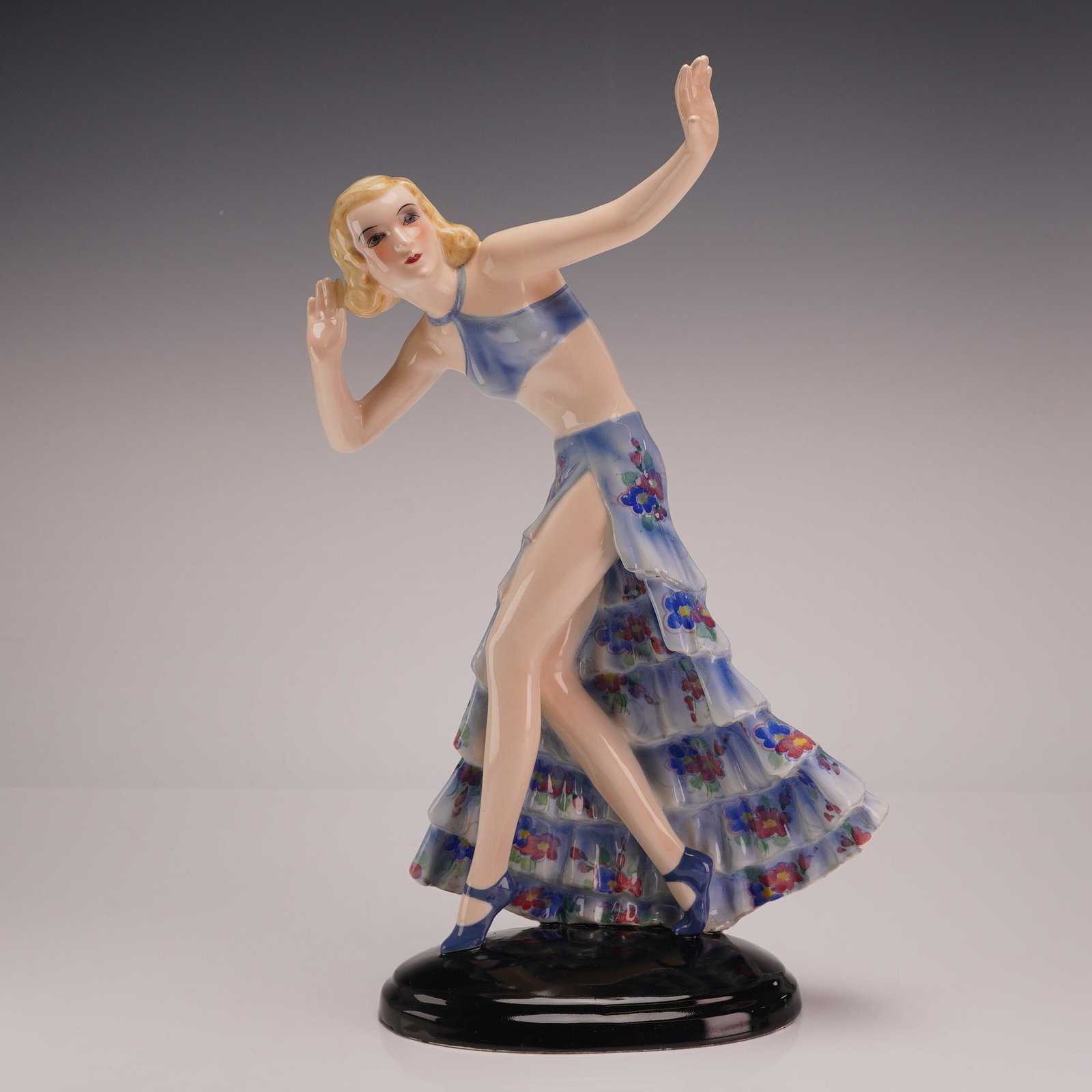 Goldscheider by Josef Lorenzl Art Deco Dancer Figurine, Model 7058: The figure depicts a blonde woman in a dynamic pose with arms raised, wearing a blue halter-style crop top and a long, tiered, ruffled skirt. The skirt features a white ground decorated with blue, red