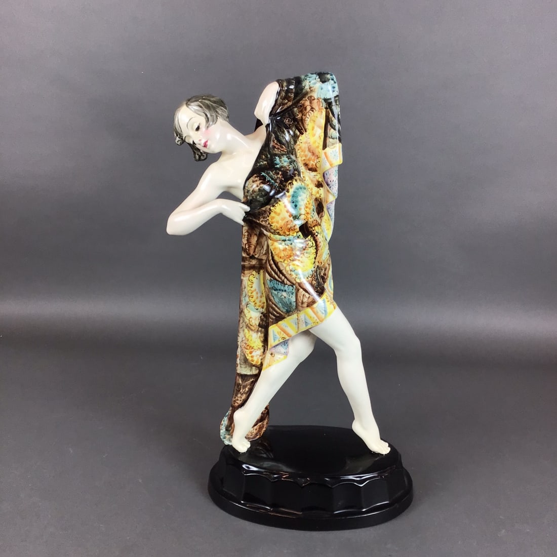Keramos Austria Sculpture Woman Dancer: This Art Deco glazed porcelain figurine depicts a dancer. The figure is captured in a dynamic pose, partially draped in a large, vibrantly patterned robe featuring intricate motifs in shades of yellow