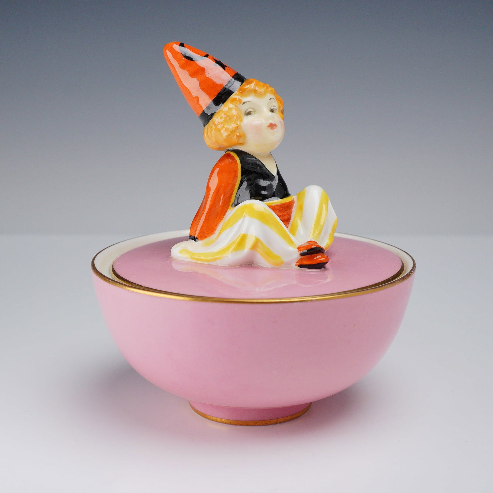 Royal Doulton Cassim Art Deco Porcelain Powder Jar: This Royal Doulton Cassim figurine powder bowl with lid is constructed of fine porcelain. The circular lidded bowl features a solid pink ground color with gold accents applied to the rims and the slig