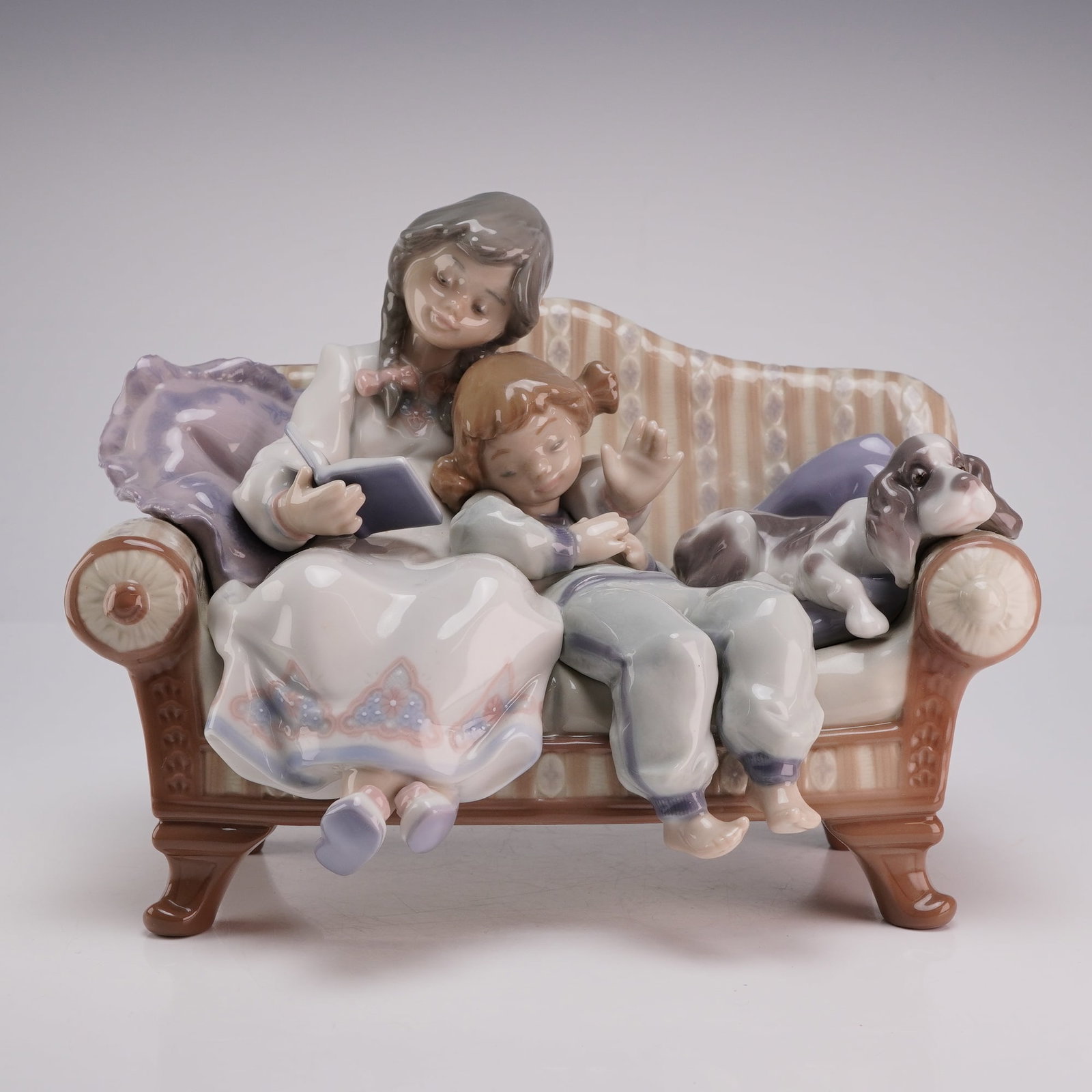 Lladro Porcelain Figurine, Big Sister 01005735: A Lladro glazed porcelain figurine titled Big Sister, model number 5735. This piece depicts two young girls seated on a striped classical-style sofa. The older girl on the left is reading from an open