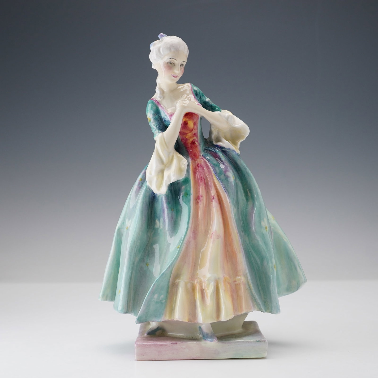 Royal Doulton Figurine, Camilla HN1711: A hand painted porcelain figure of a lady in 18th-century attire. She stands upon a square base and clasping her hands at her chest. She wears a flowing gown in soft green and aqua tones with floral a