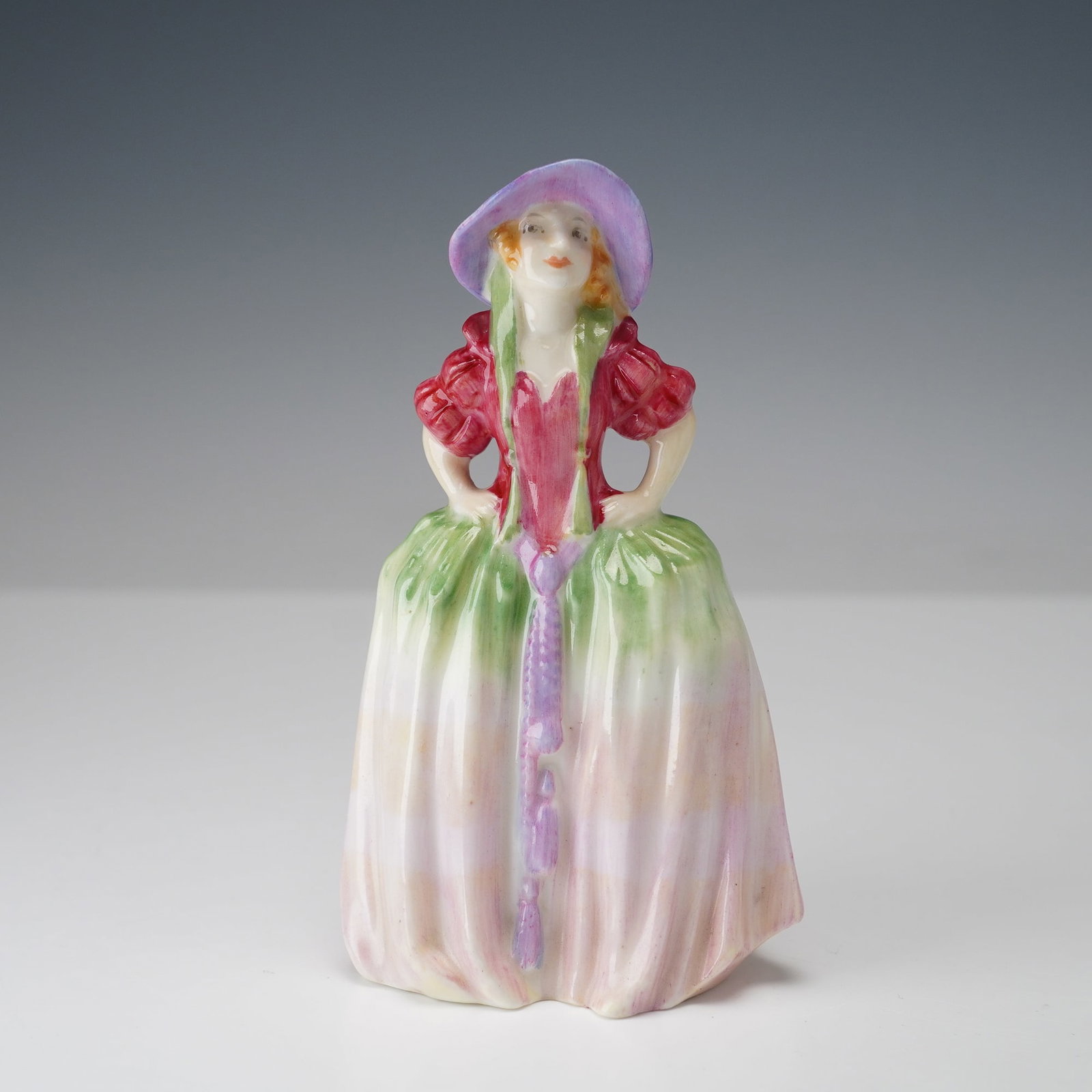 Royal Doulton Mini Figurine, Patricia M7: A Royal Doulton miniature figurine, Patricia M7. The figure depicts a young woman in a pink and green gown with puffed sleeves and a lavender hat, hands on hips in a confident pose. Hand-painted in so