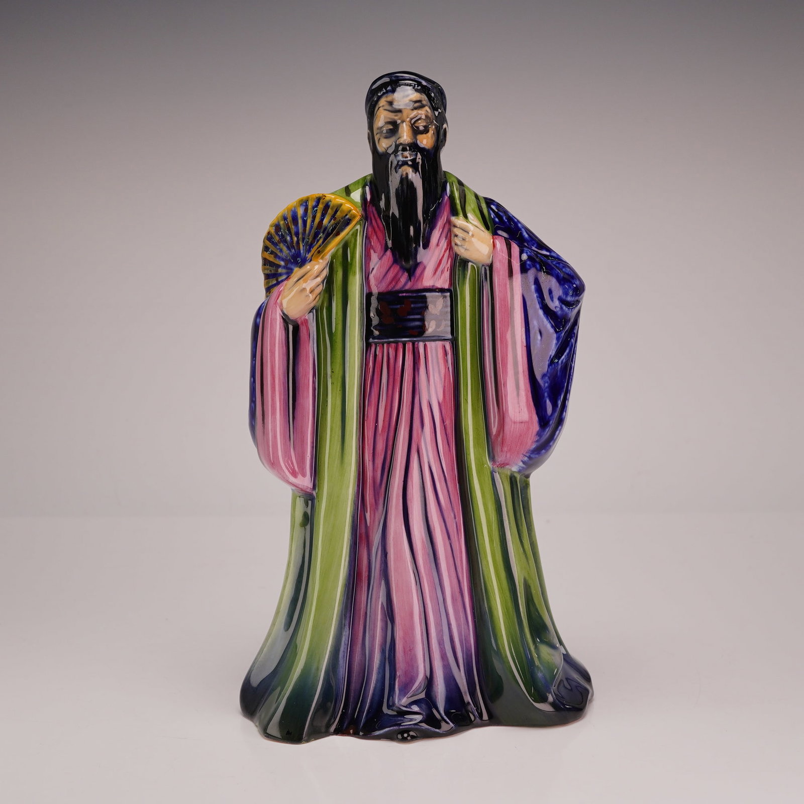 Royal Doulton Prototype Figurine, Confucius: The figure is depicted in traditional Chinese scholarly attire, wearing a long pink under-robe with a black sash, a green outer cloak, and a blue shawl. He holds a yellow and blue folding fan in his r