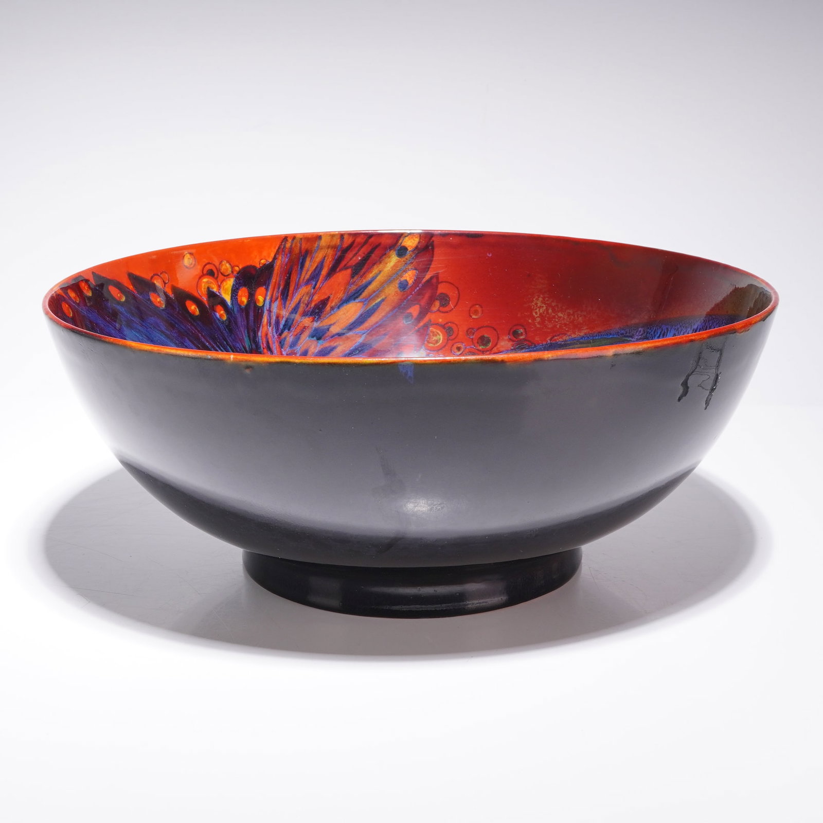 Royal Doulton by Charles Noke ,Rare Large Flambe Sung Phoenix Bowl: The interior is decorated with a stylized phoenix in vibrant shades of cobalt blue, royal purple, and orange. The bird is set against a mottled background of cream, yellow, and red flambe glazes, acce