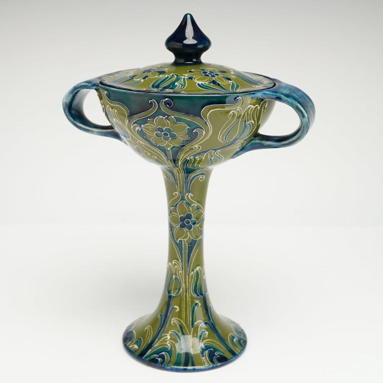 James Macintyre by William Moorcroft Florian Ware Lidded Bonbonniere: This vessel features a slender stem rising from a widely flaring circular foot, with two prominent stylized handles framing a broad rim. The body and lid are finished in a green and blue ground, adorn