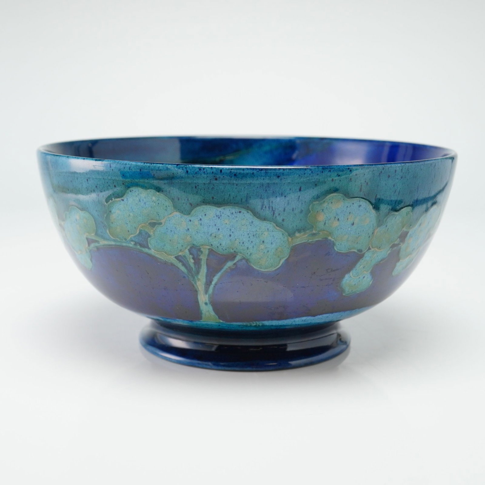 Moorcroft Pottery Footed Bowl, Moonlit Blue: The deep, circular form features a slightly flared rim and is decorated using a hand-painted, tube-lined technique. The landscape design depicts stylized trees rendered in varying hues of cobalt, turq