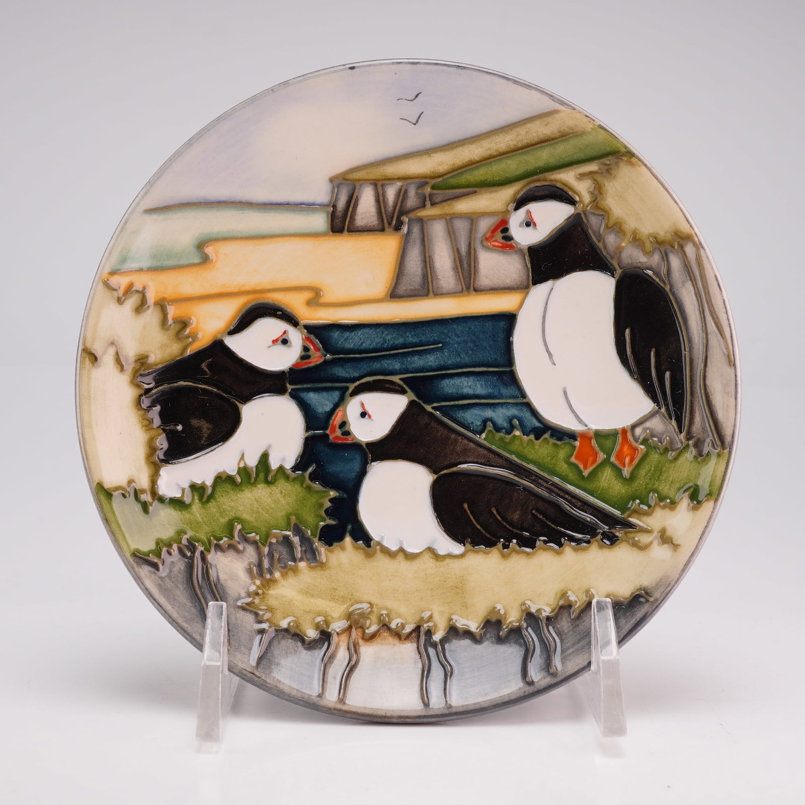 Moorcroft Pottery by Carole Lovatt Pin Dish, Puffins: The circular dish is decorated using the tube-lining technique with hand-painted polychrome glazes. The scene depicts three puffins on a rocky shoreline against a background of sea, cliffs, and a pale