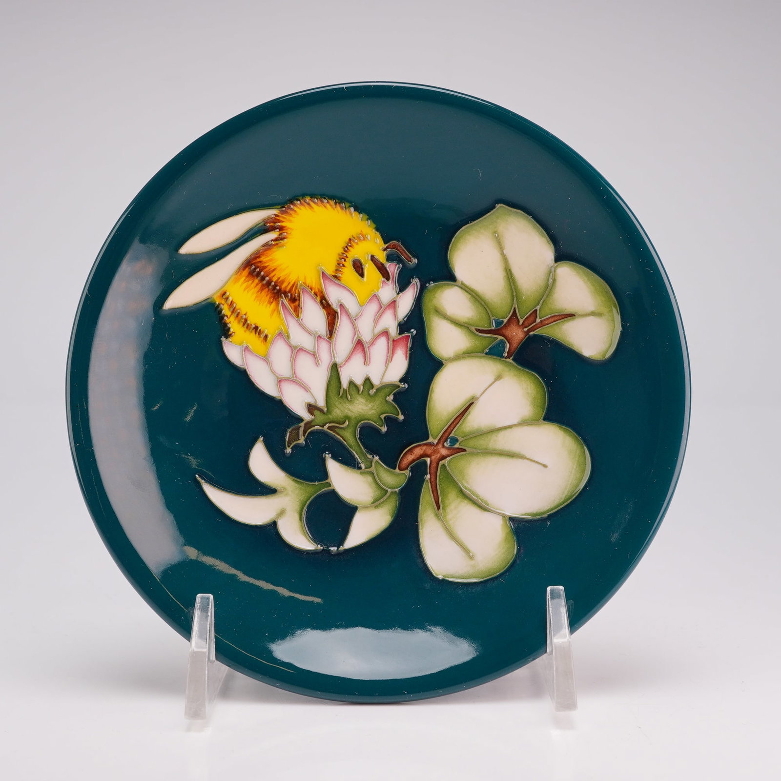 Moorcroft Pottery by Philip Richardson Pin Dish, Bumblebee: The central motif depicts a yellow and black bumblebee landing on a white flower with pink-tipped petals, accompanied by green trilobed leaves. The decoration is set against a deep teal glaze. The und