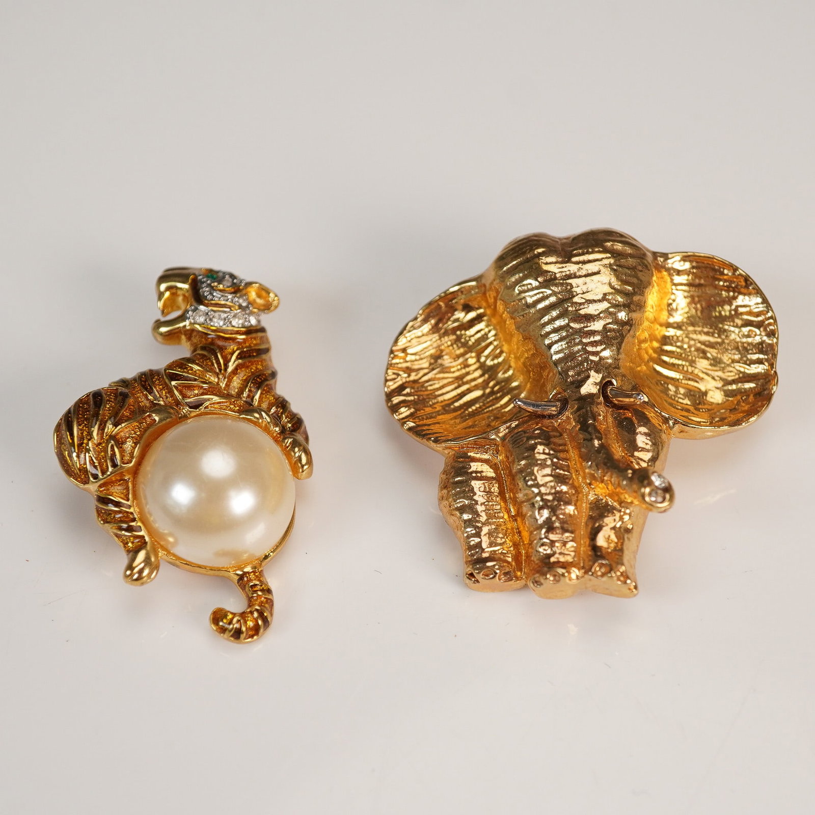 2pc Gold Tone Animal Brooch Set, Landau Tiger and Elephant: Lot of two whimsical animal-themed brooches featuring a gold tone Landau tiger brooch adorned with a faux pearl belly and pave rhinestone accents, and an elephant head brooch with textured detailing a