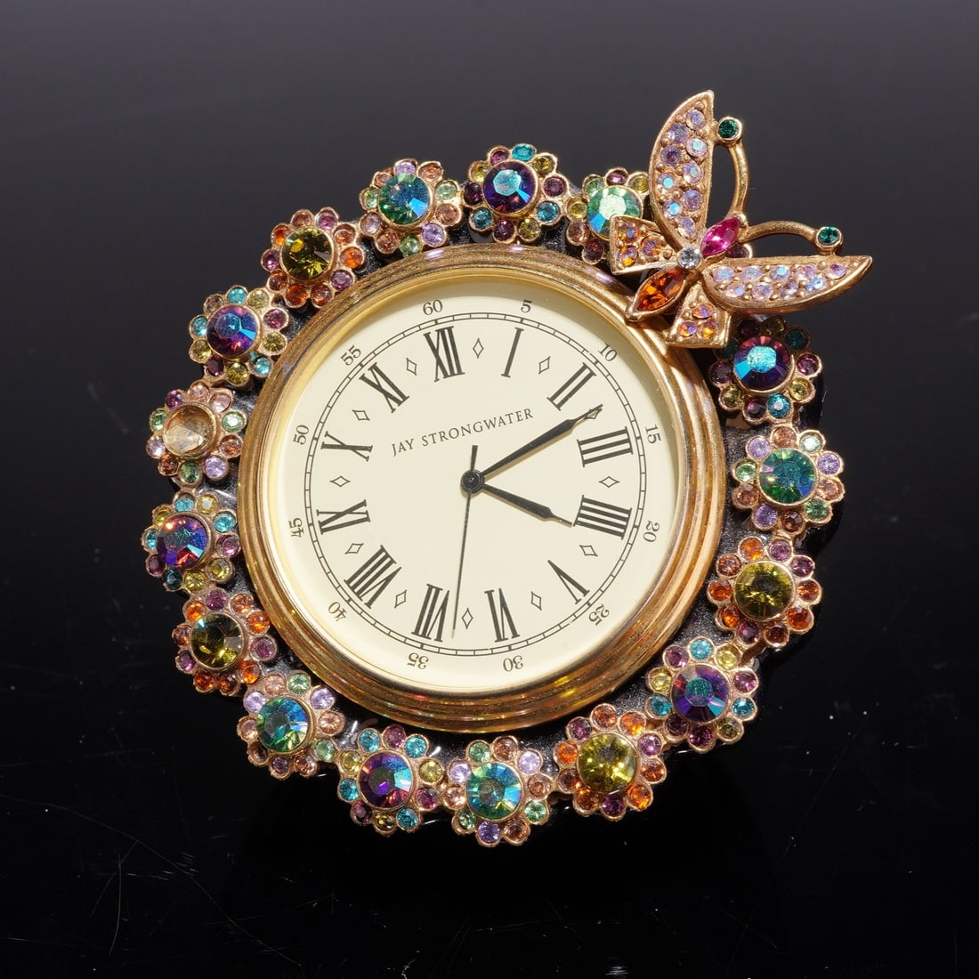 Jay Strongwater Flower Edge Jeweled Desk Mantel Clock w/ Butterfly: This Jay Strongwater flower edge jeweled desk or mantel clock with butterfly is constructed with a cast metal frame in a gold-tone finish. The circular frame is decorated with an array of multi-colore