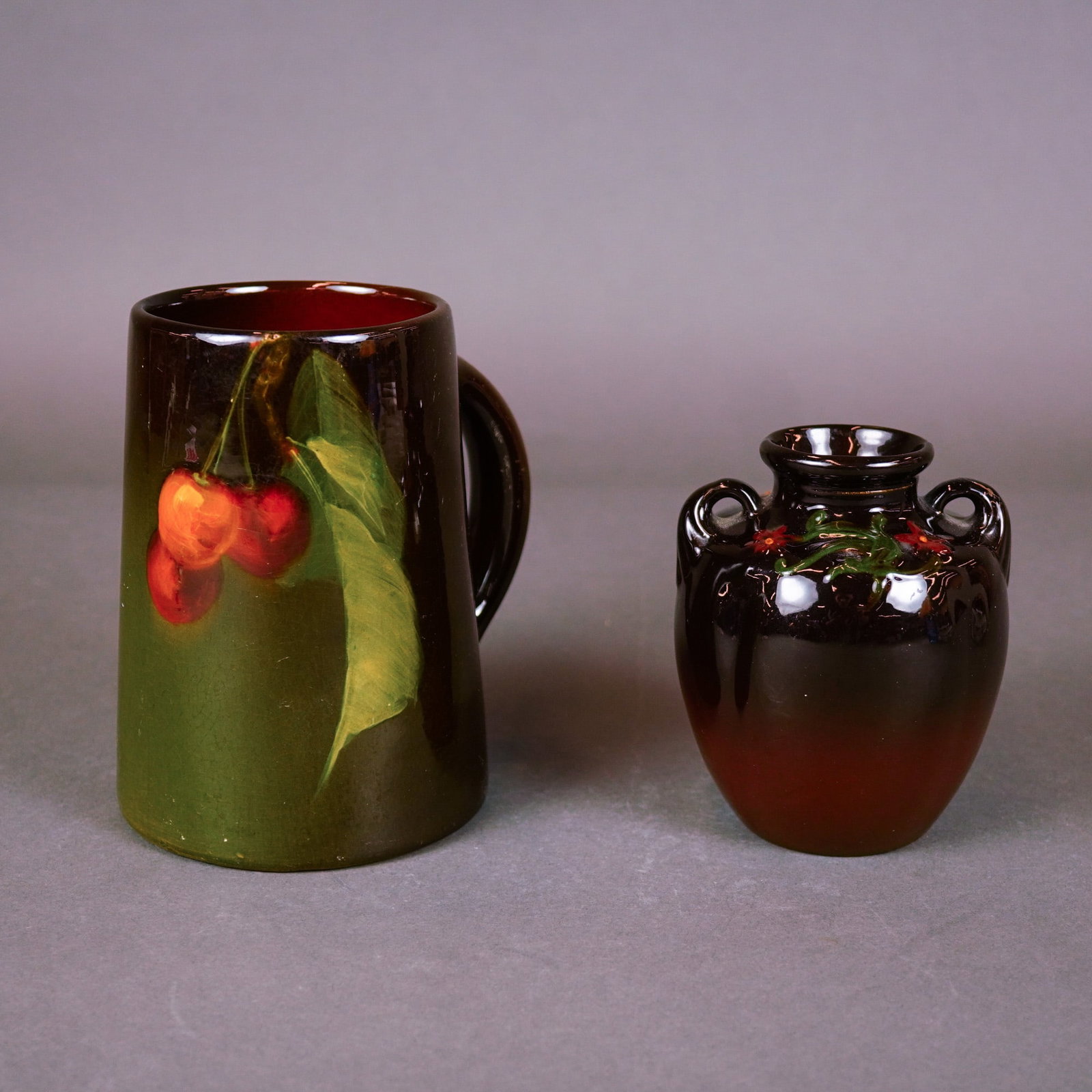 Weller Pottery Louwelsa Cherries Mug and Floretta Floral Vase: A pair of early American art pottery pieces from Weller Pottery. The first is a Louwelsa earthenware mug featuring a hand-painted underglaze slip decoration of cherries on a stem with a large green