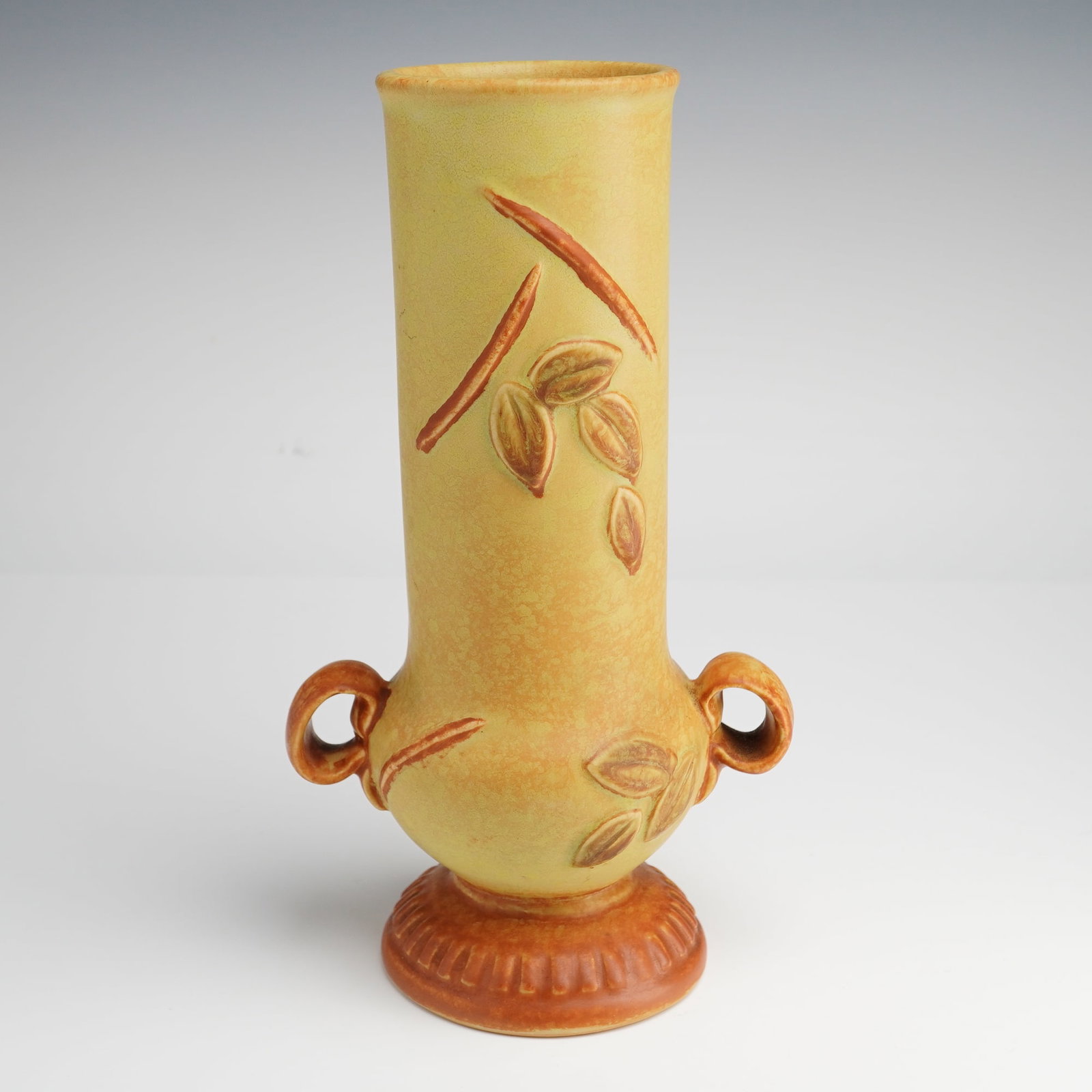 Weller Pottery Golden Glow Blossom and Leaves Handled Vase: This American art pottery earthenware vase was produced by Weller Pottery as part of the Golden Glow line, circa 1930. The vessel features a tall cylindrical form with a slightly bulbous lower body