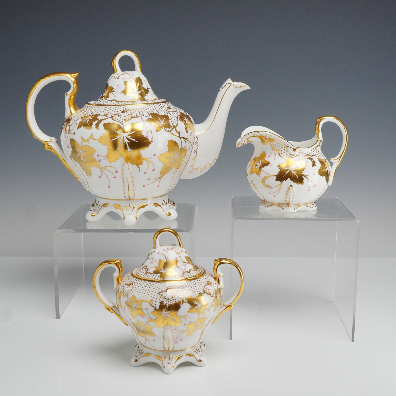 3pc Royal Chelsea Gilded Bone China Tea Service - Teapot, Creamer and Lidded Sugar Bowl: A trio of Royal Chelsea English bone china tea service items comprising a teapot, a double-handled sugar bowl, and a creamer. Each piece features a matching ornate gilded leaf and vine pattern on a