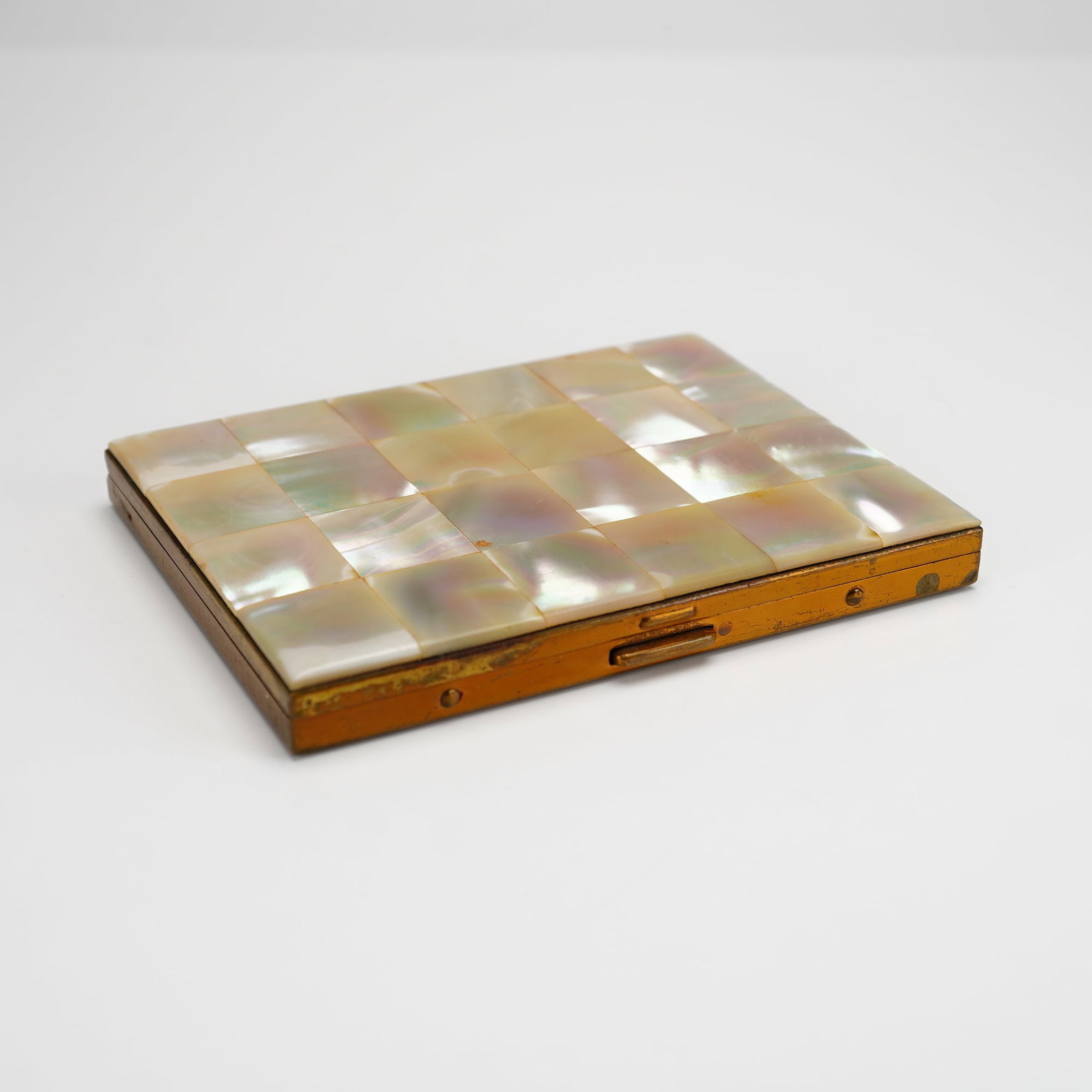 Mother of Pearl and Brass Compact Case: This rectangular compact case features a decorative checkerboard inlay of iridescent mother-of-pearl tiles on the front cover, set within a brass frame. The reverse of the case consists of a plain bra