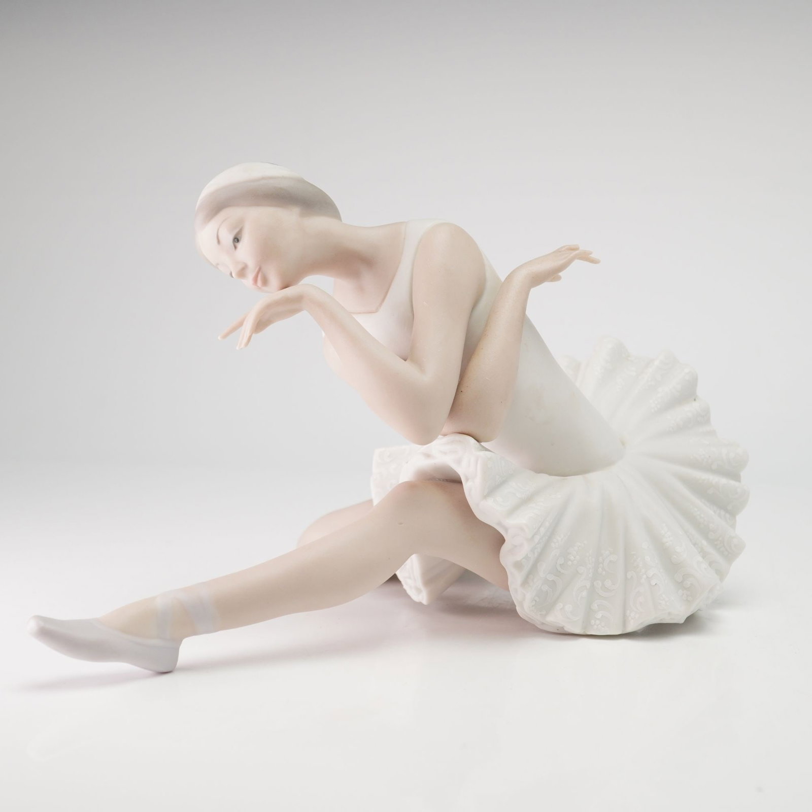 Lladro Matte Porcelain Figurine Death of the Swan 01014855: This Lladro matte porcelain figurine depicts a seated ballerina in a contemplative pose. The figure is dressed in a classic white tutu featuring an embossed scrolling foliate and floral pattern, paire