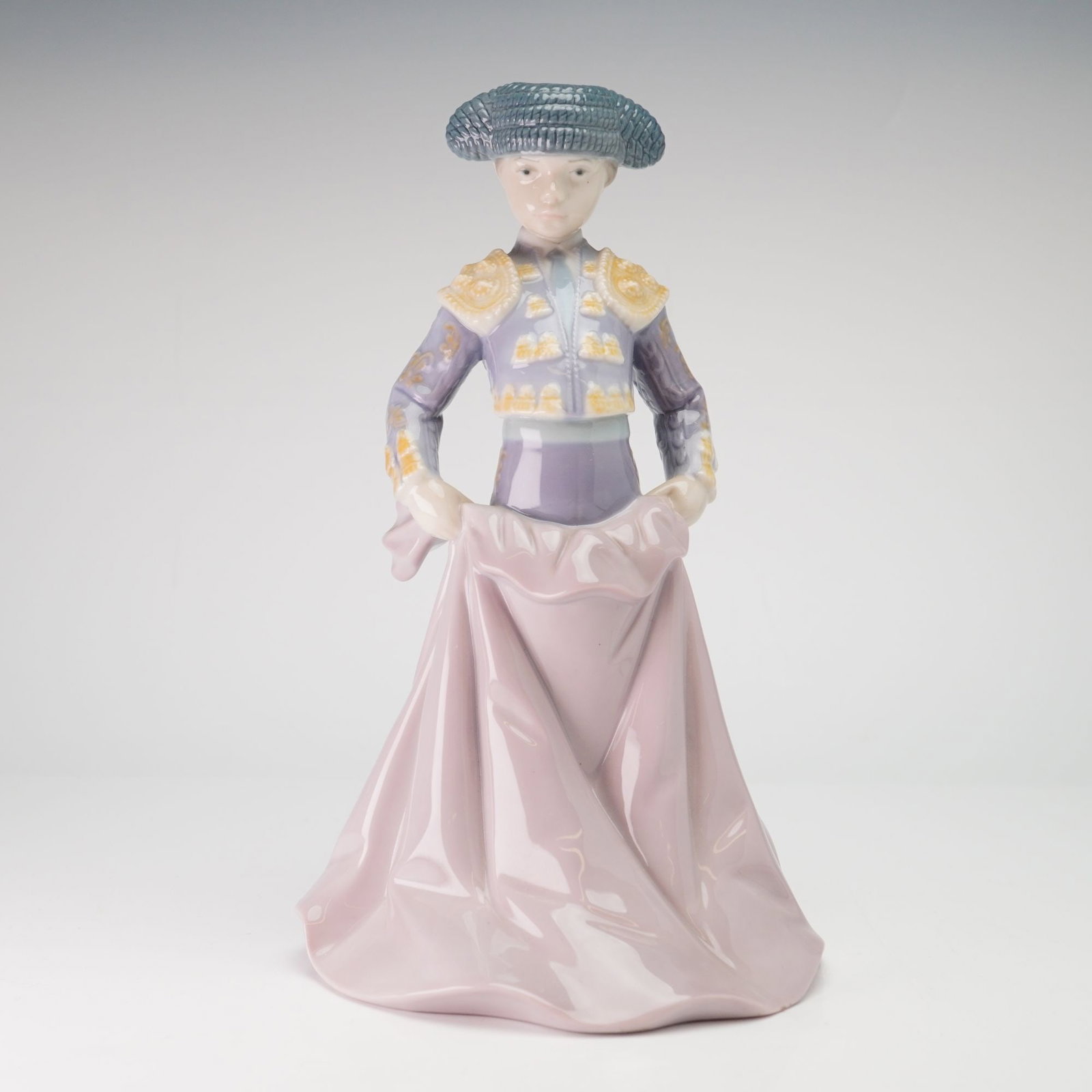 Lladro Porcelain Figurine Young Torero 01006438: The figure depicts a young bullfighter in a traditional suit of lights, or traje de luces, featuring a lavender and purple color scheme with gold-tone decorative elements. The torero wears a blue grey