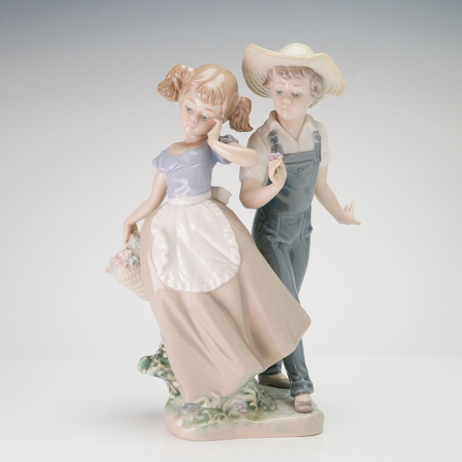 Lladro Porcelain Figurine Love in Bloom 01005292: The scene depicts a young boy and girl in pastoral attire. The boy is modeled in a walking pose wearing a white collared shirt, blue denim-style overalls with crossed suspenders, and a wide-brimmed st