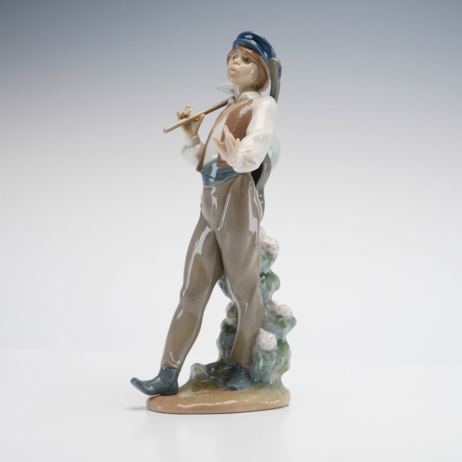 Lladro Porcelain Figurine Wandering Minstrel 01005676: The figure depicts a young boy in mid-stride, dressed in a blue cap, a brown vest over a long-sleeved white shirt, a blue sash, and brown trousers tucked into blue shoes. He carries a wooden walking s