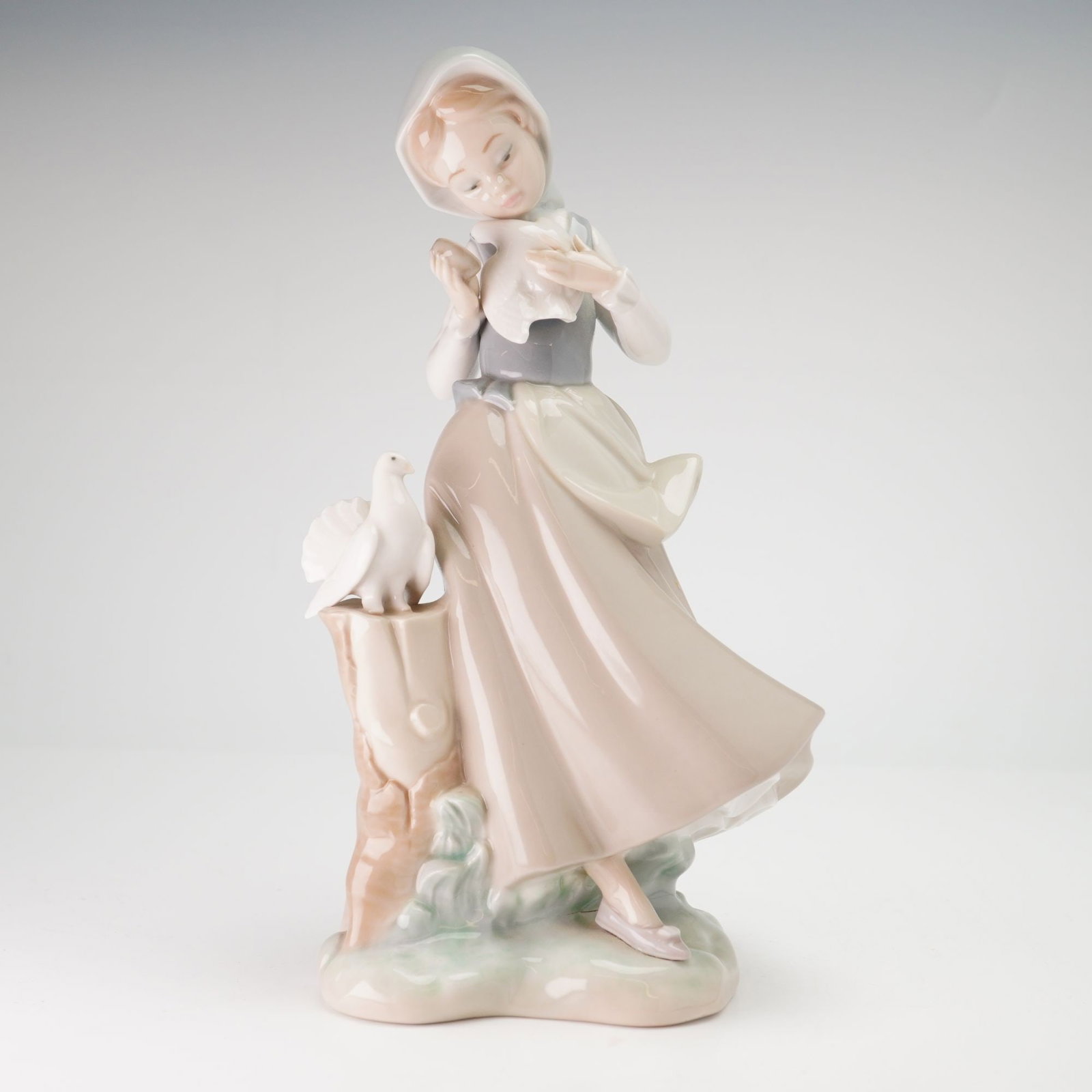 Lladro Porcelain Figurine, Girl with Pigeons 01004915: The piece depicts a young girl in traditional rustic attire, featuring a white headscarf, a gray bodice, and a flowing tan skirt with a cream-colored apron. She is positioned next to a tree stump, hol