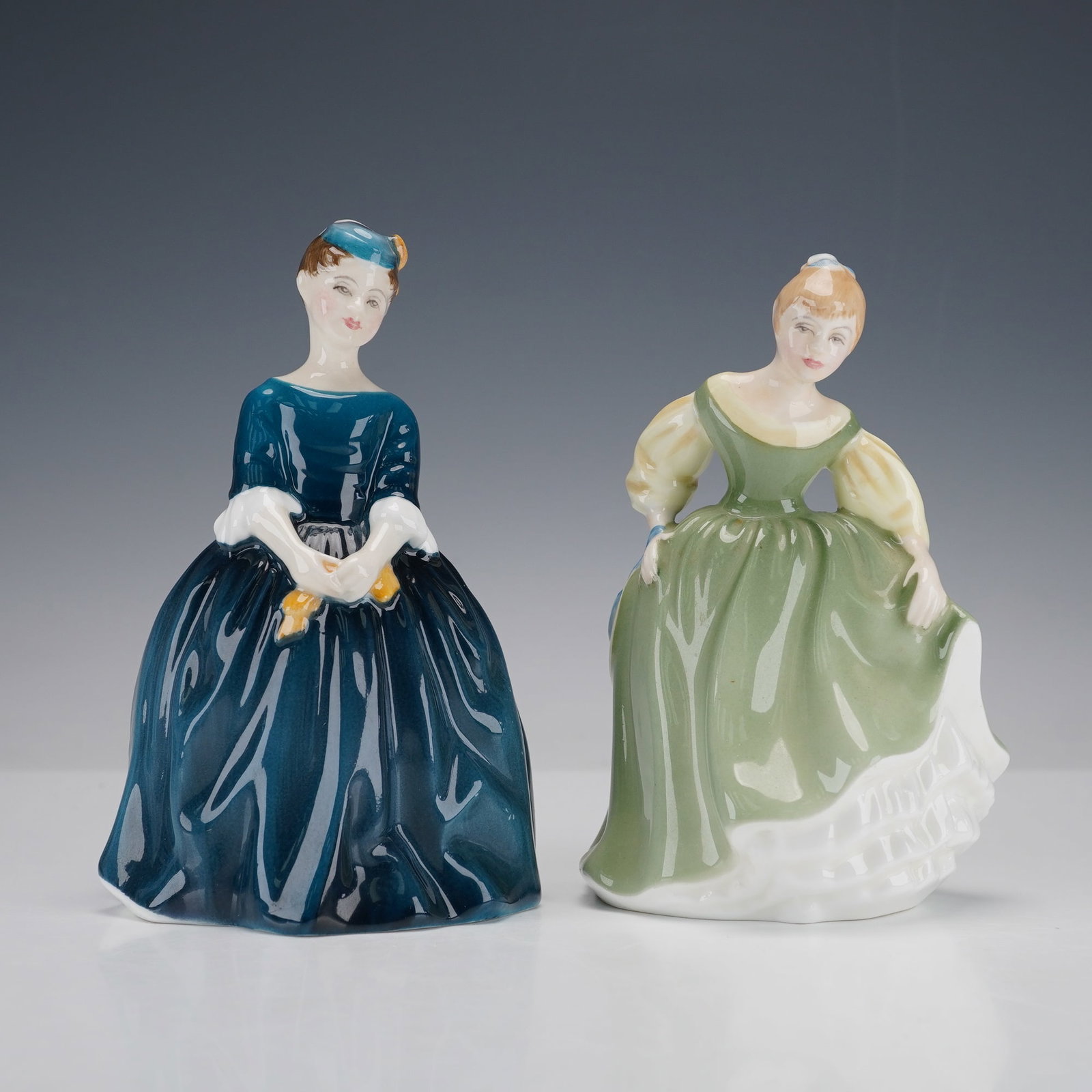 2pc Royal Doulton Figurines, Cherie HN2341 and Fair Maiden HN2211: A graceful duo of finely detailed figurines including Cherie, wearing a deep blue gown and matching hat adorned with a yellow ribbon, and Fair Maiden, dressed in a flowing green dress with pale accent