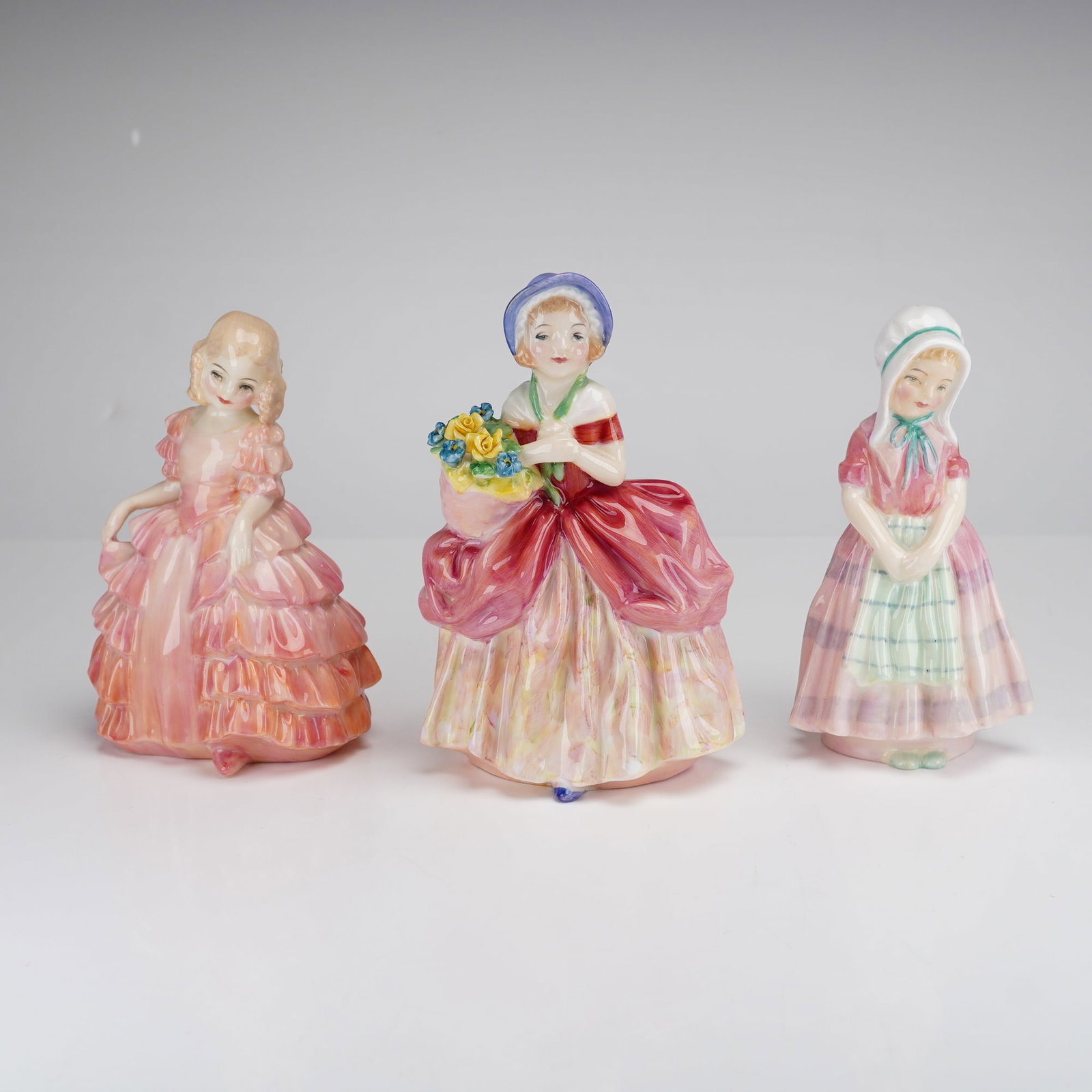 Royal Doulton, Three Figurines: Rose, Cissie, and Tootles: This collection features three vintage Royal Doulton glazed bone china figurines, each portraying a young girl in traditional attire. The first, Rose (HN 1368), depicts a girl in a multi-tiered pink a